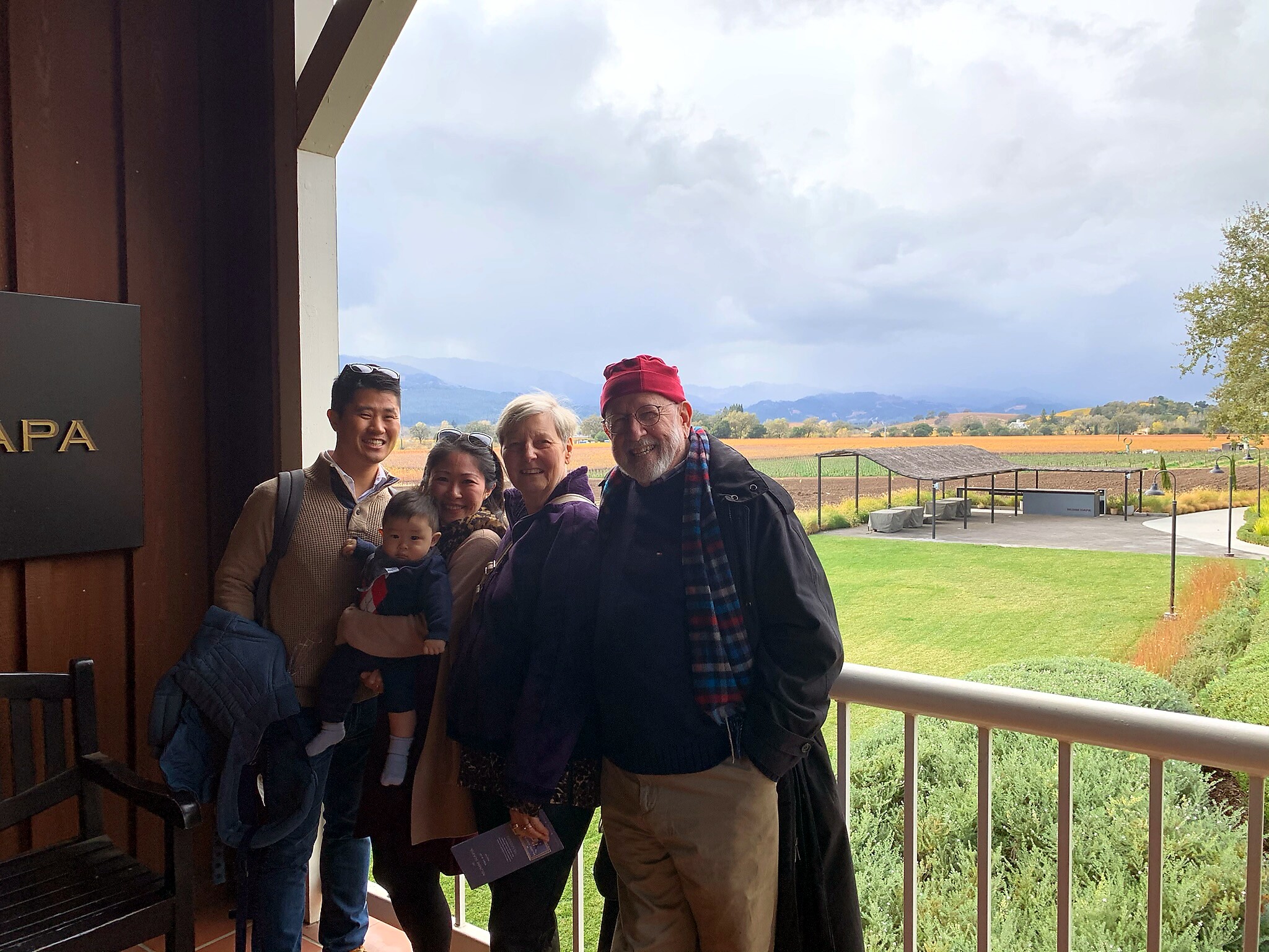 Group of five people posing on a covered outdoor balcony with a baby, smiling, and a rural landscape of fields and hills behind them.