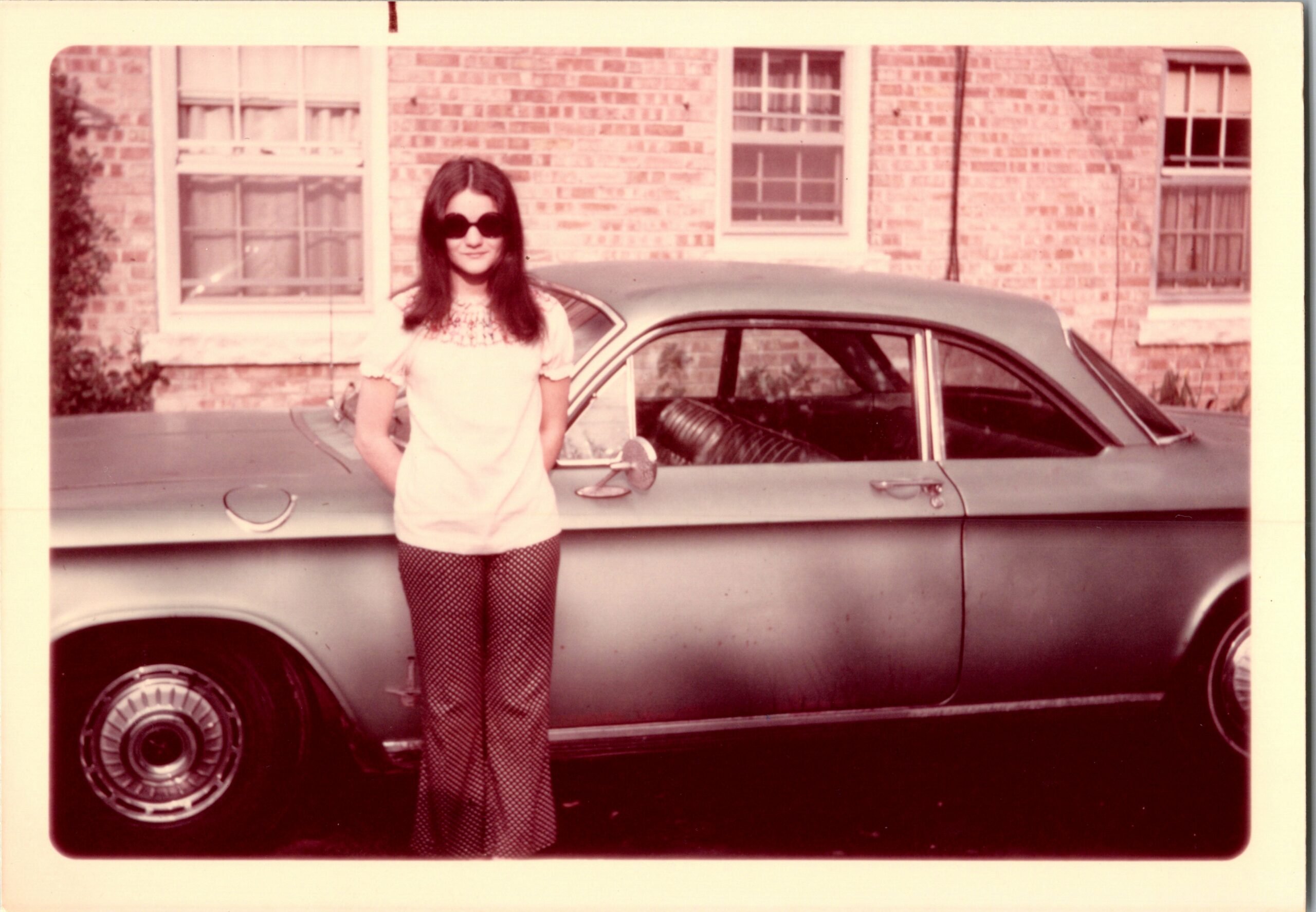 Young girl wearing sunglasses, a white embroidered top, and polka-dot pants stands beside a vintage car in a brick-building courtyard.