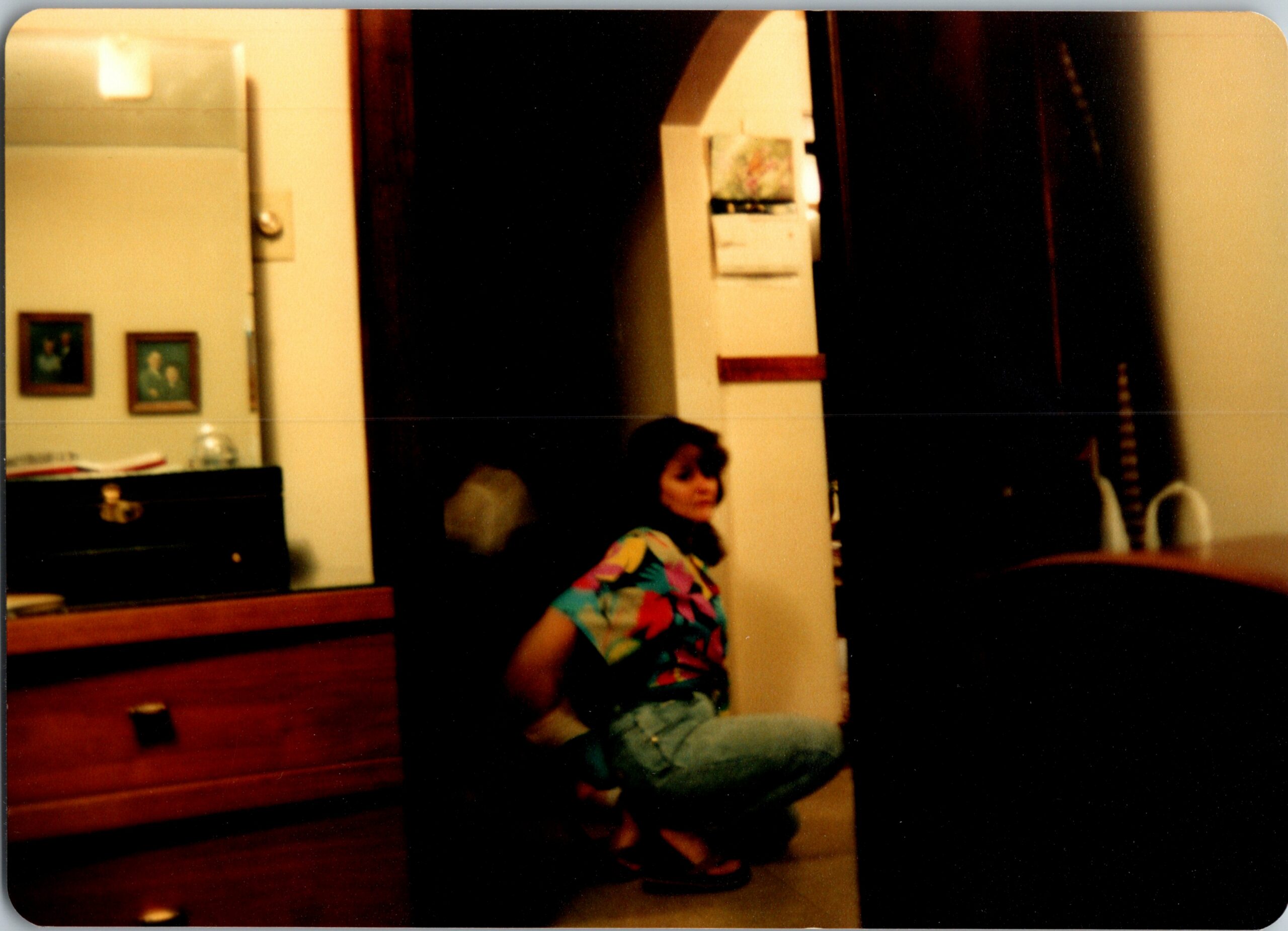 Woman in a bright floral top sitting in a living room next to a white pillar, with a piano and framed photos on the wall behind her.