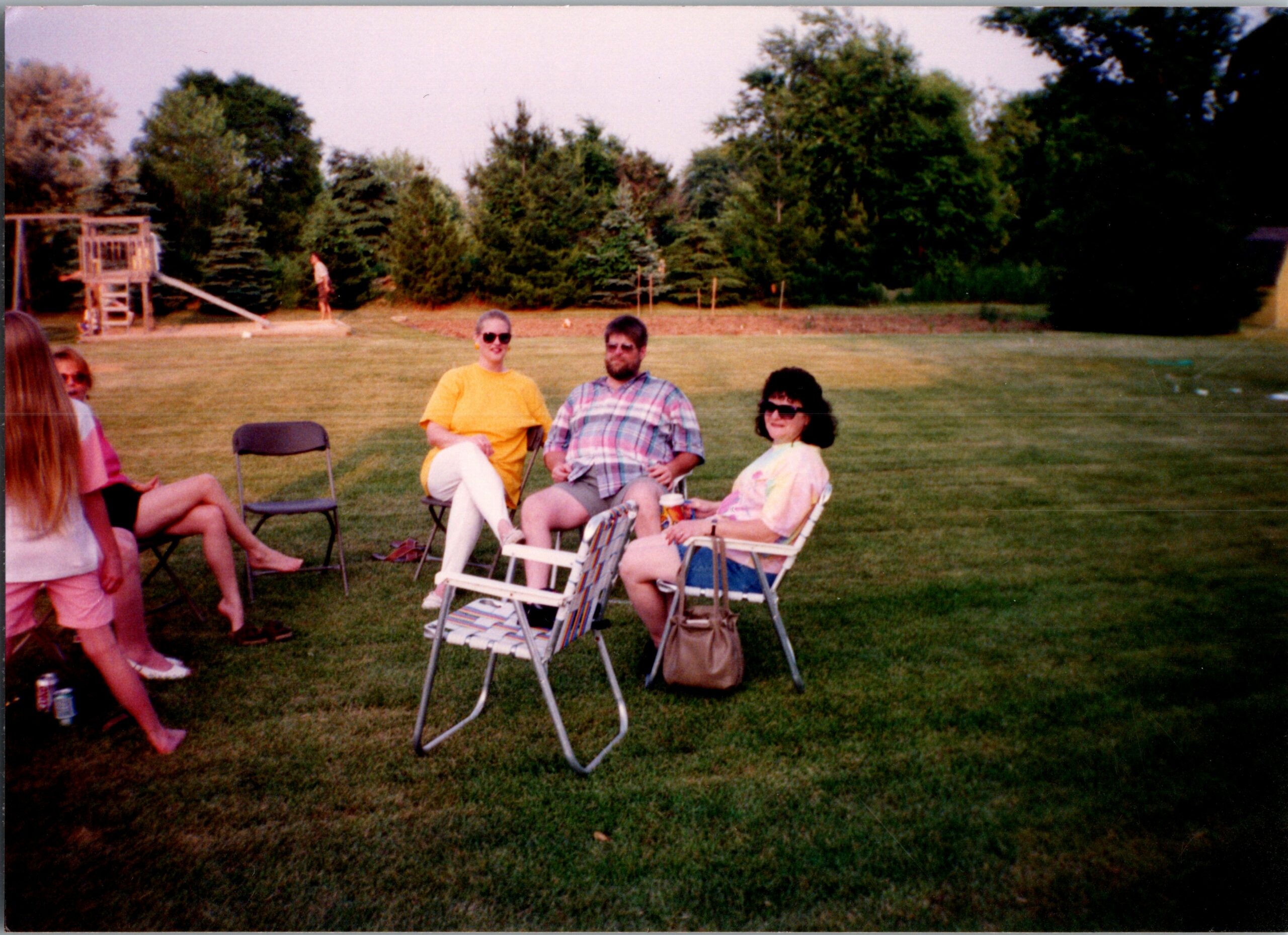 Group of people seated on lawn chairs in a backyard, with a playset and trees in the background.