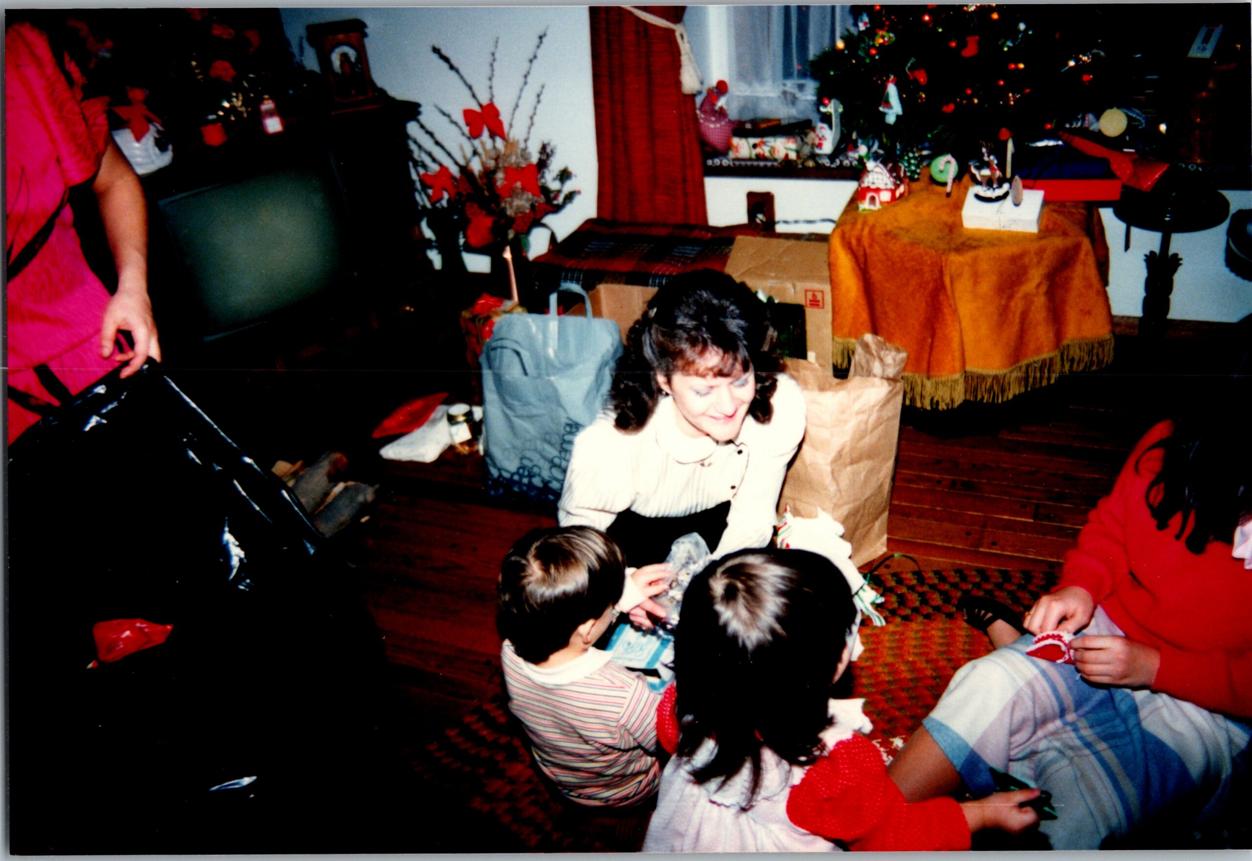 Children gathered on a living room floor opening gifts, with a decorated Christmas tree and festive decorations in the background.