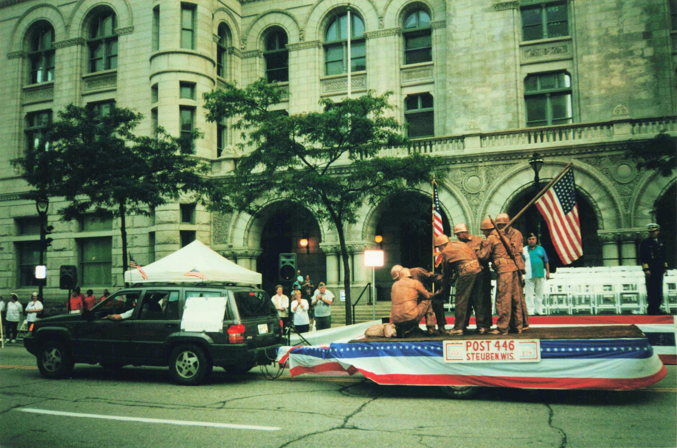 Parade scene with flag-bearing uniformed workers on a float in front of a historic stone building; spectators line the street behind.
