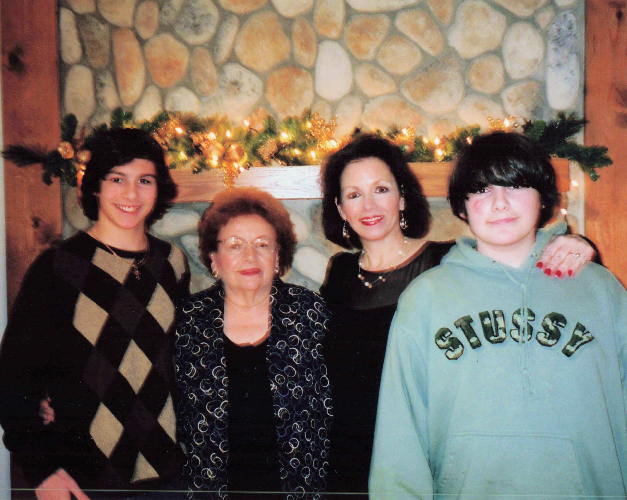 Group of smiling women posing for a holiday photo in front of a stone fireplace with garland and lights.