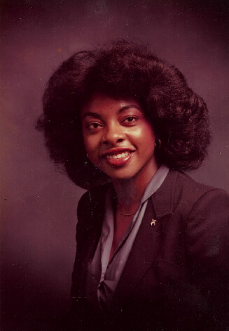 Studio portrait of a smiling Black woman with voluminous hair, wearing a dark blazer and light blouse.