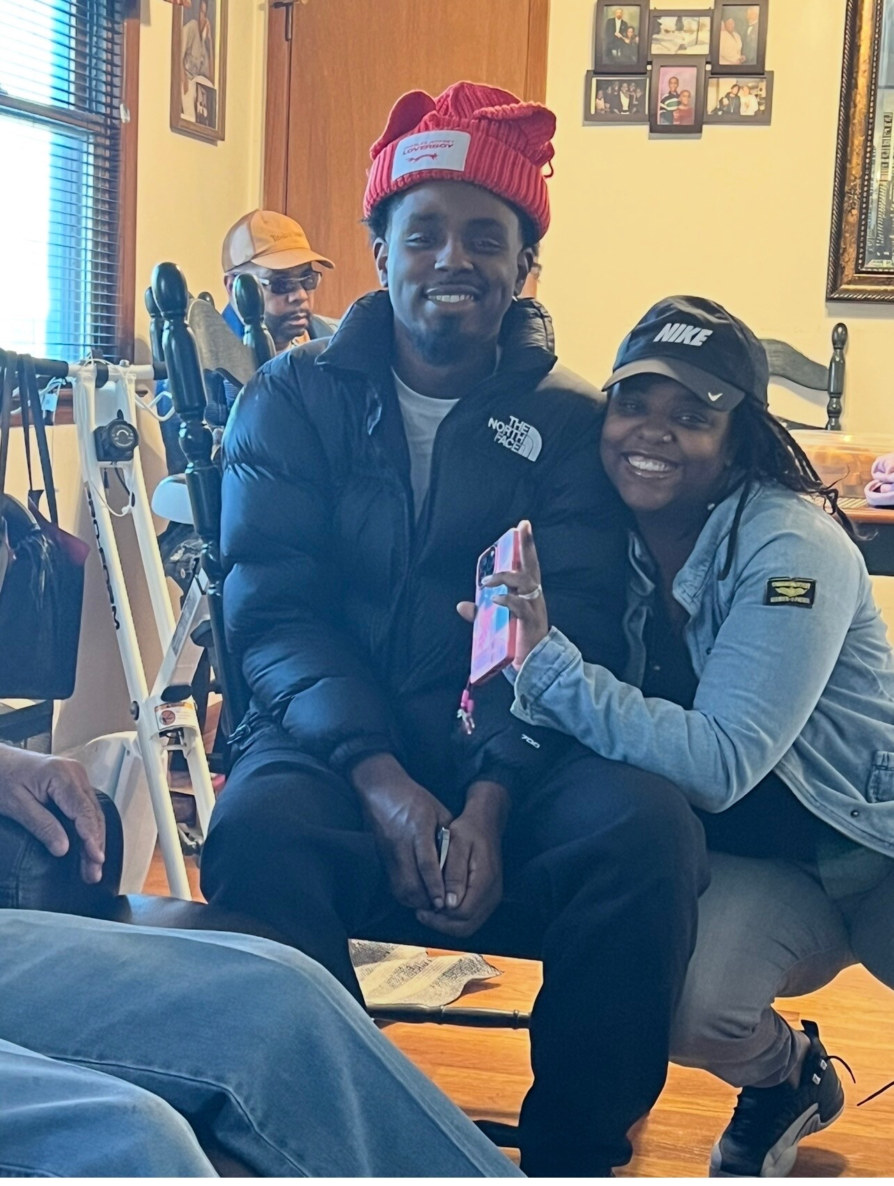 Two smiling adults posing indoors together; man in a red knit hat and black puffer jacket, woman wearing a black Nike cap and gray jacket beside him.