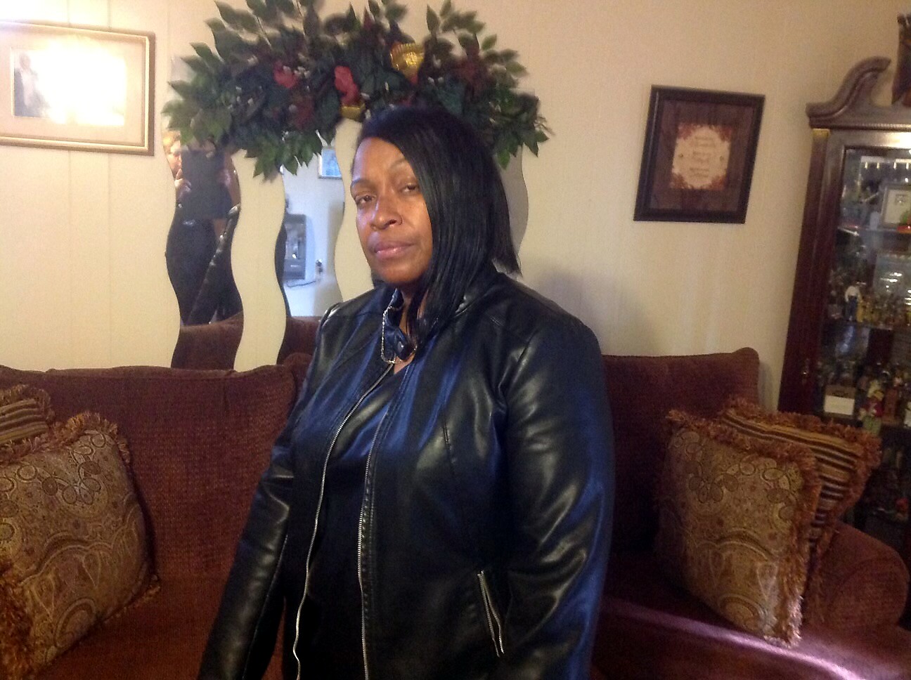 Woman wearing a black leather jacket stands beside a brown sofa in a warmly lit living room with decorative pillows and plants behind her.