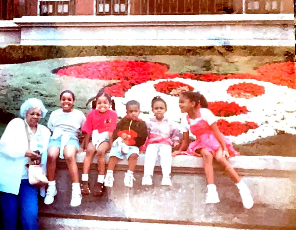 An elderly woman with blue hair sits beside six children on a stone ledge in a garden with red and white flowers behind them.