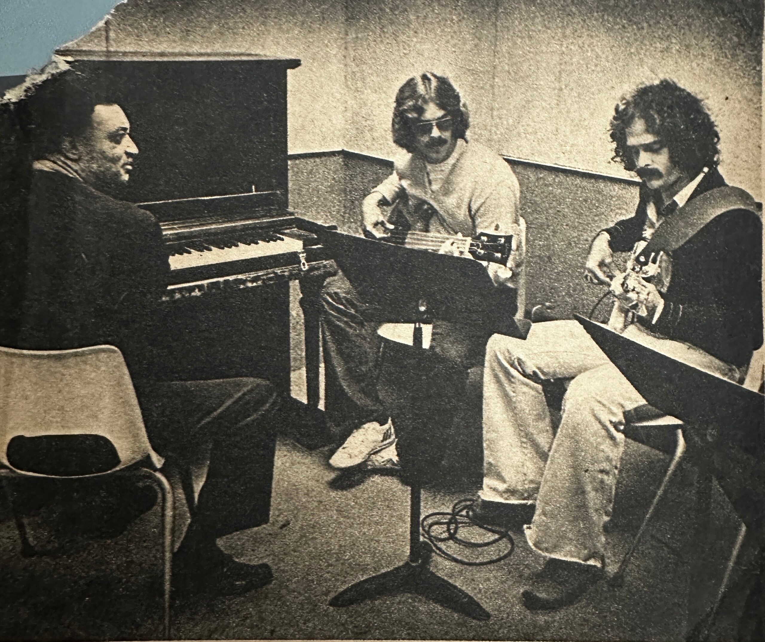 Three musicians performing: a pianist on the left and two guitarists on the right, in a black-and-white photo.