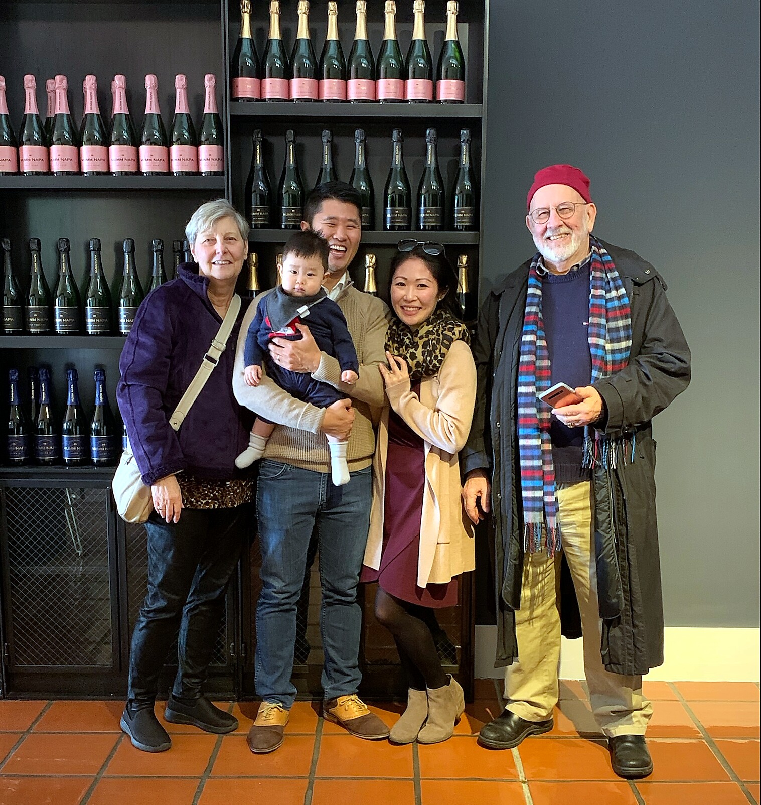 Smiling group of five adults and a baby posing in front of wine bottles on shelves in a shop.