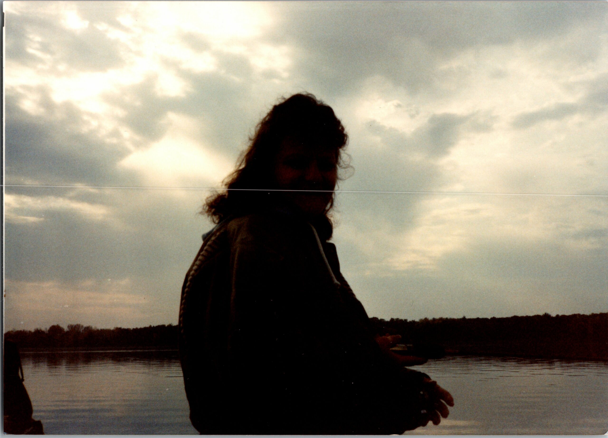 Silhouette of a person with curly hair standing by a body of water under a cloudy sky.