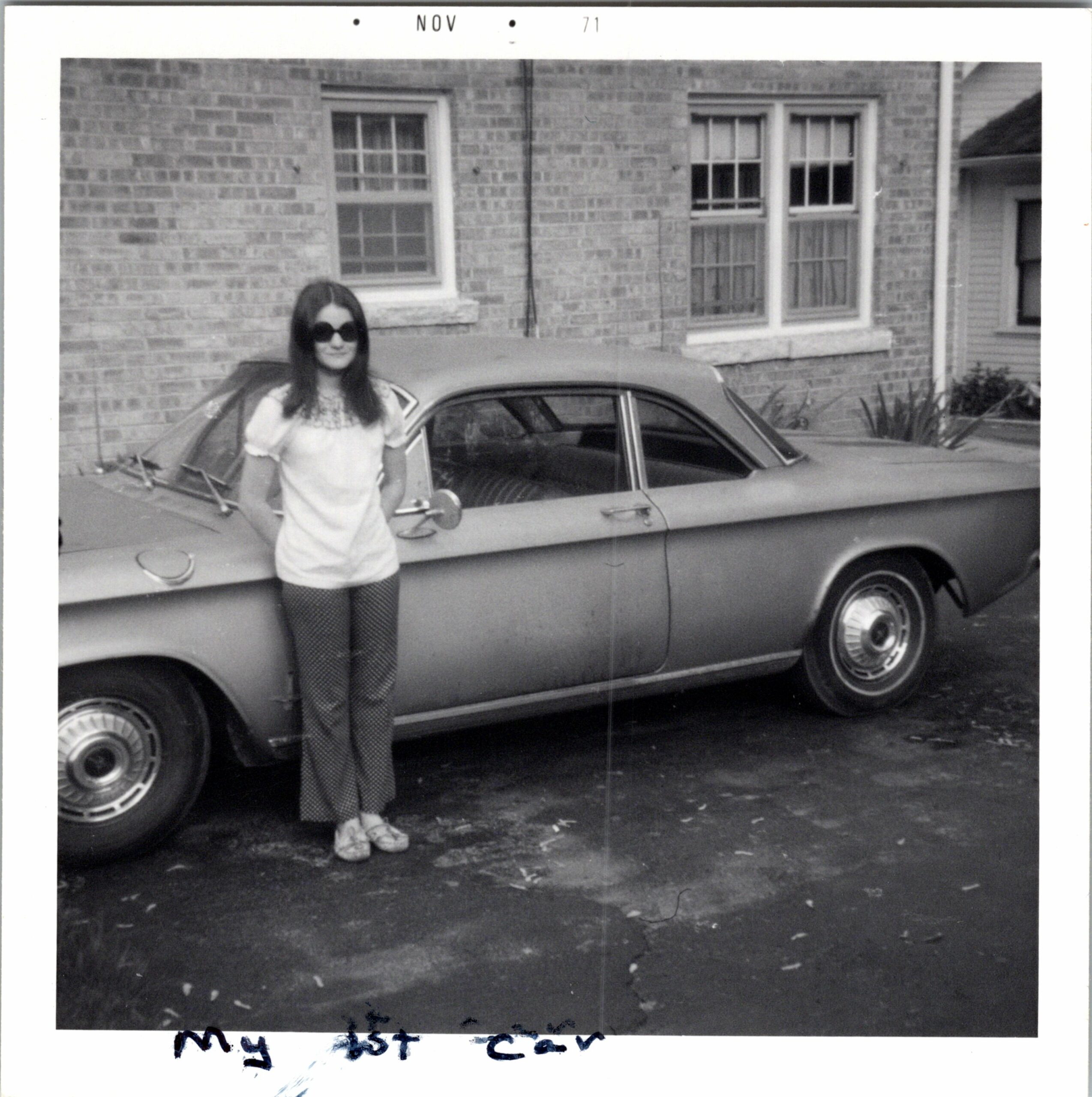 Young woman wearing sunglasses standing beside a vintage car in a driveway, brick house with windows in the background (black-and-white photo).