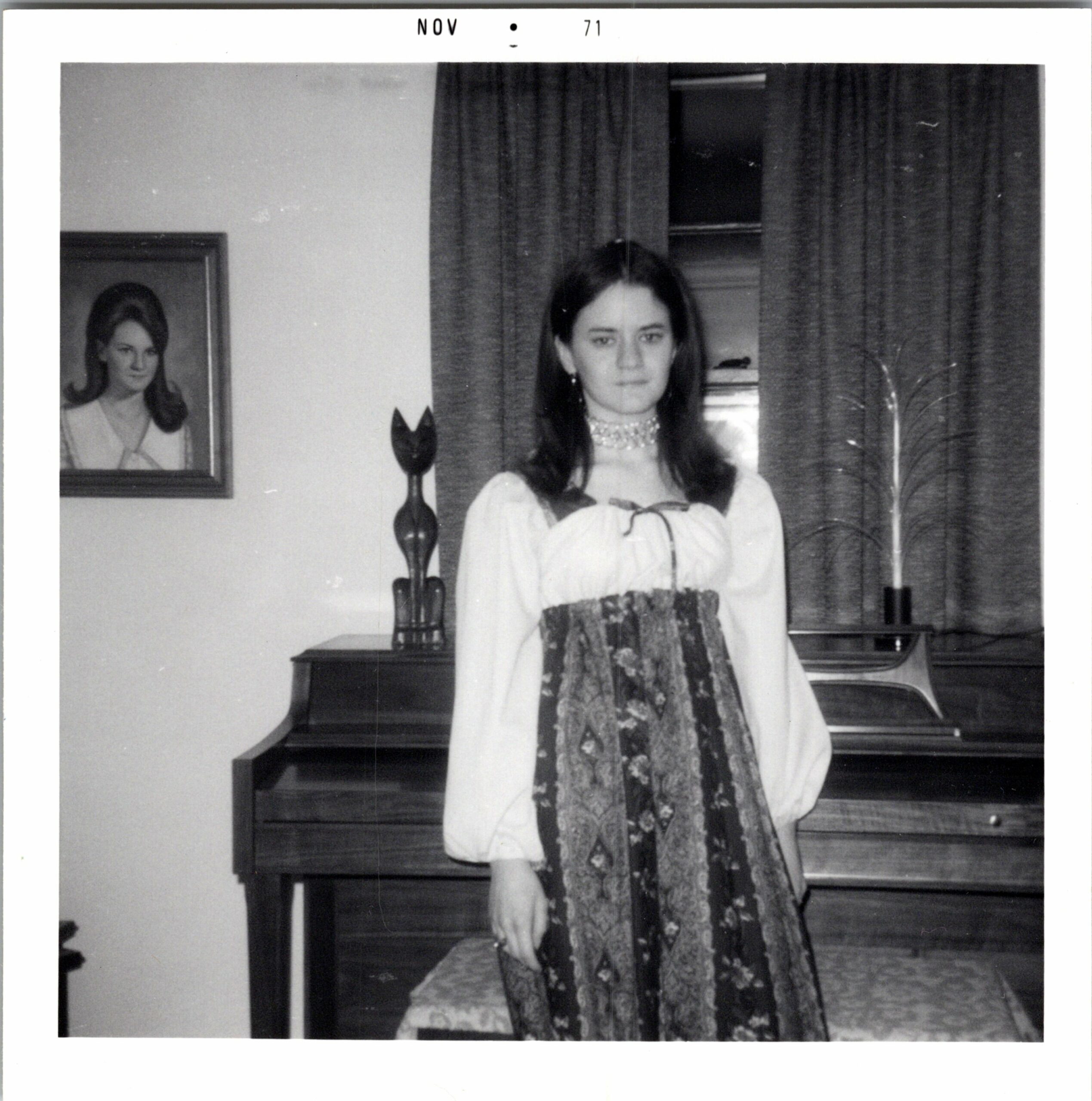 Young woman in a white blouse and ornate sash stands beside a piano in a room, with a framed portrait on the wall to her left.