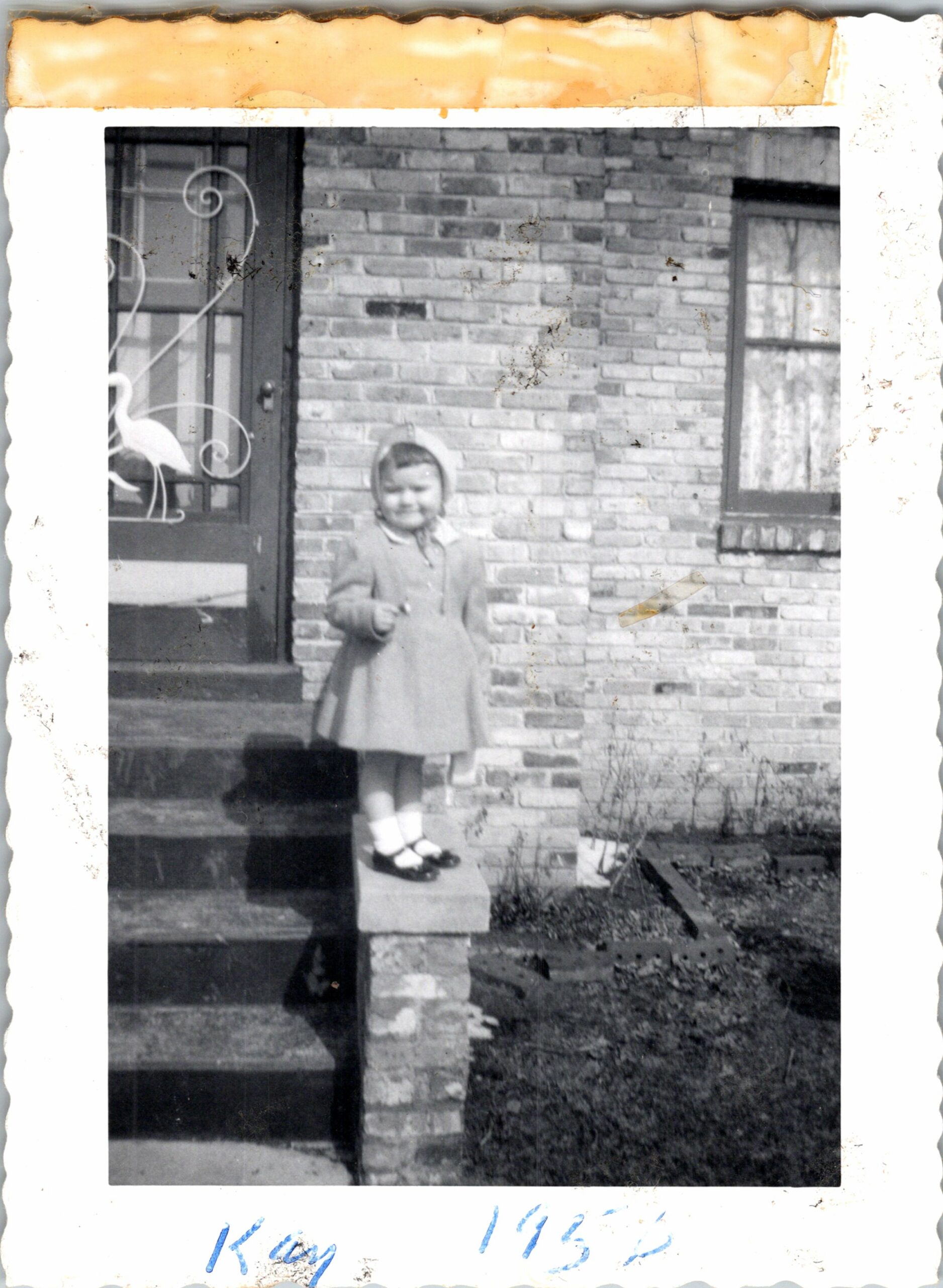 Small child in a coat and bonnet standing on brick porch steps in front of a brick house.