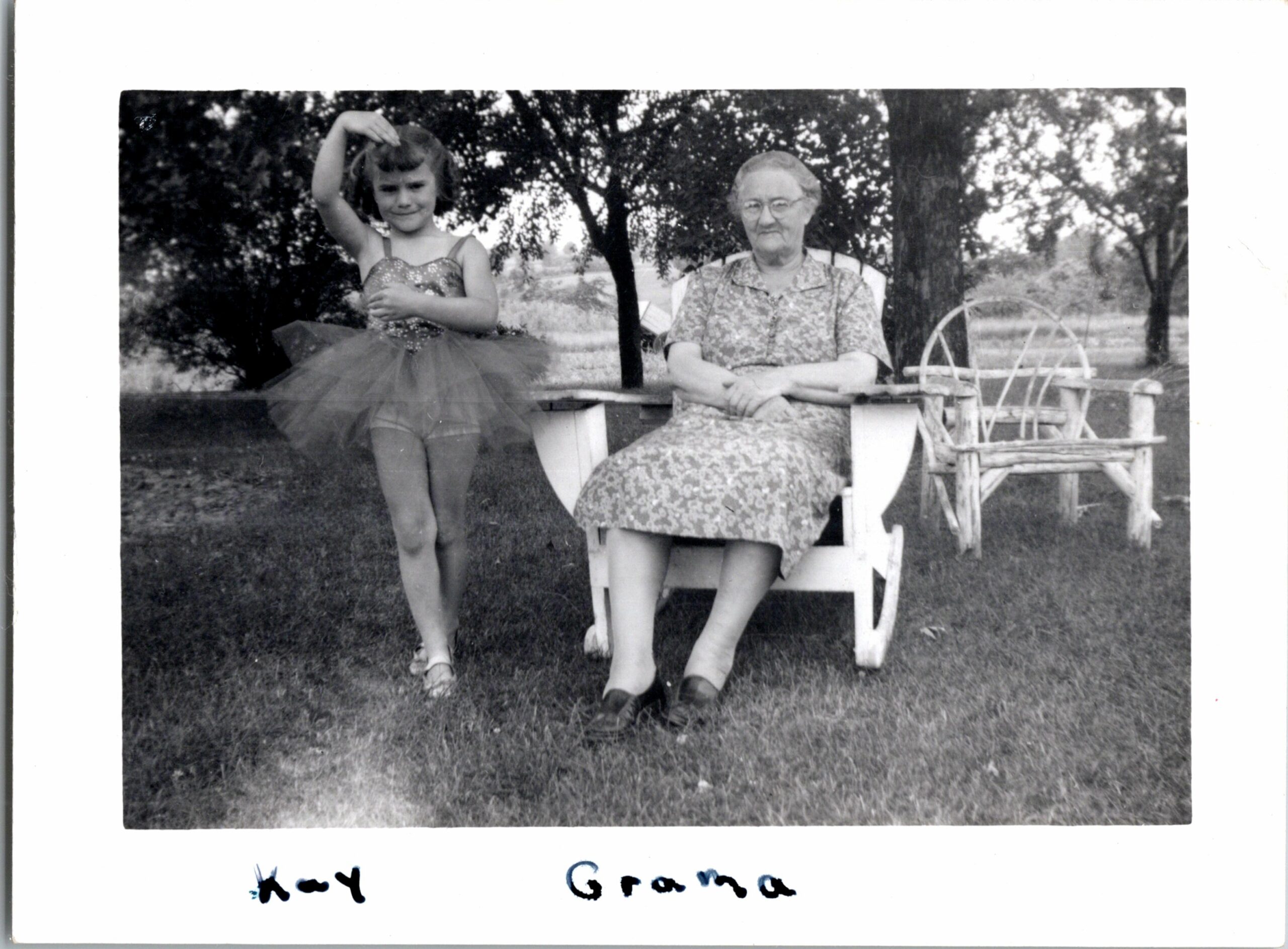 Young girl in a sparkly tutu posing beside her seated grandmother outdoors in a yard with trees behind them.