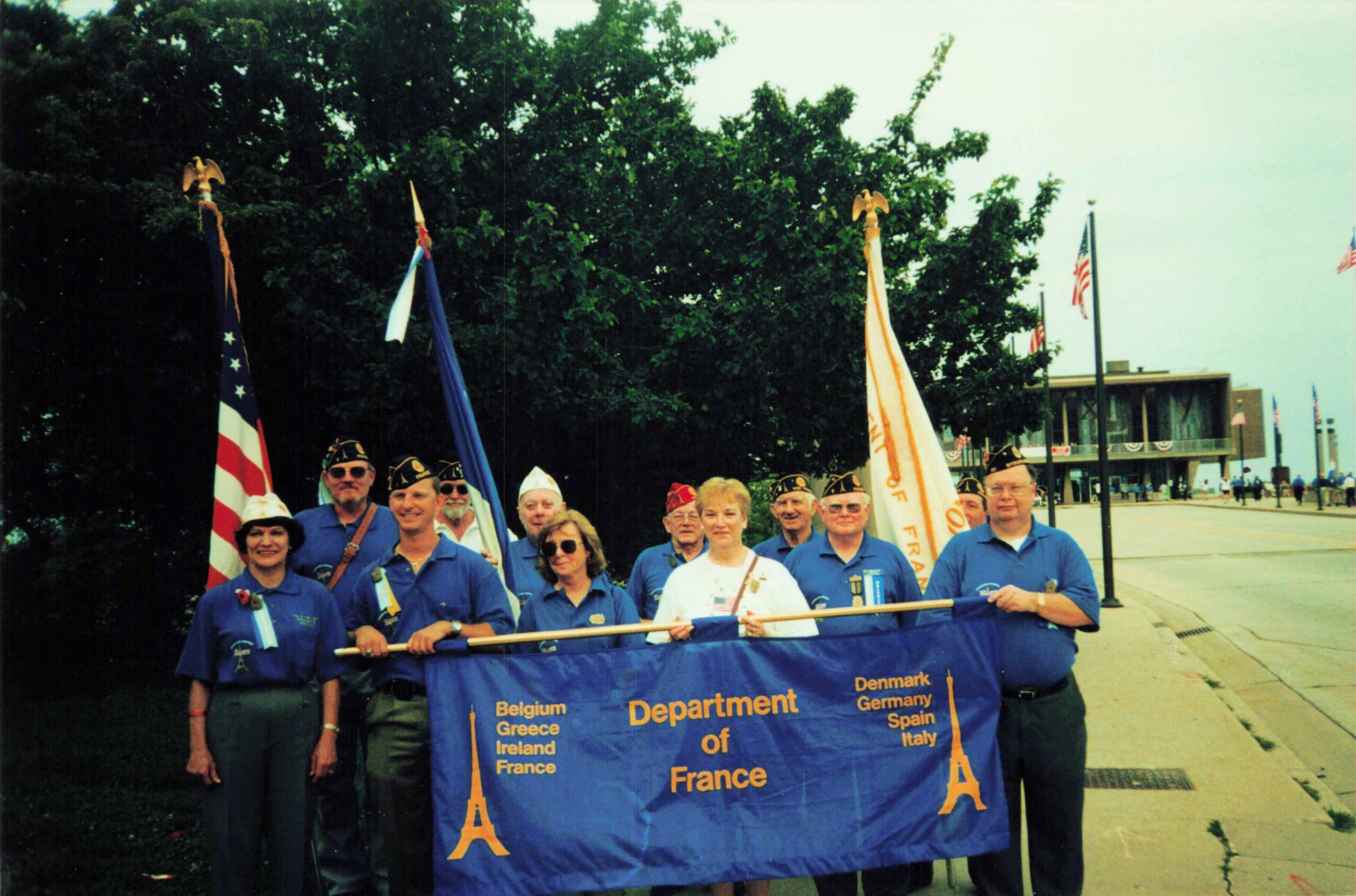 Group of veterans in blue shirts holding a blue banner listing countries (Belgium, Greece, Ireland, France) at a parade, with American flags in the background.