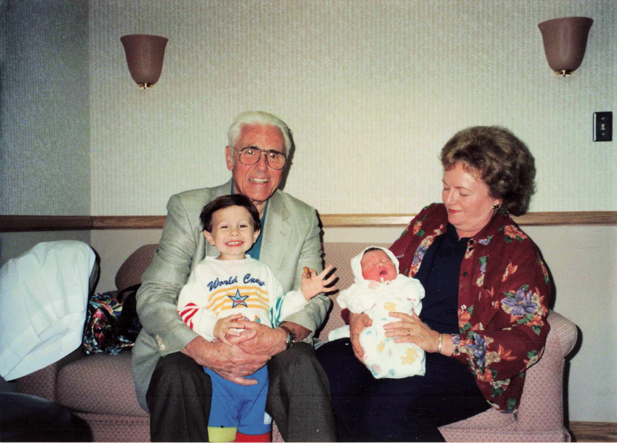Grandfather and grandmother sit on a sofa with two young children, smiling for a family photo in a living room.