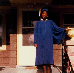 Graduate in a blue cap and gown standing on the front porch steps, smiling for the photo.