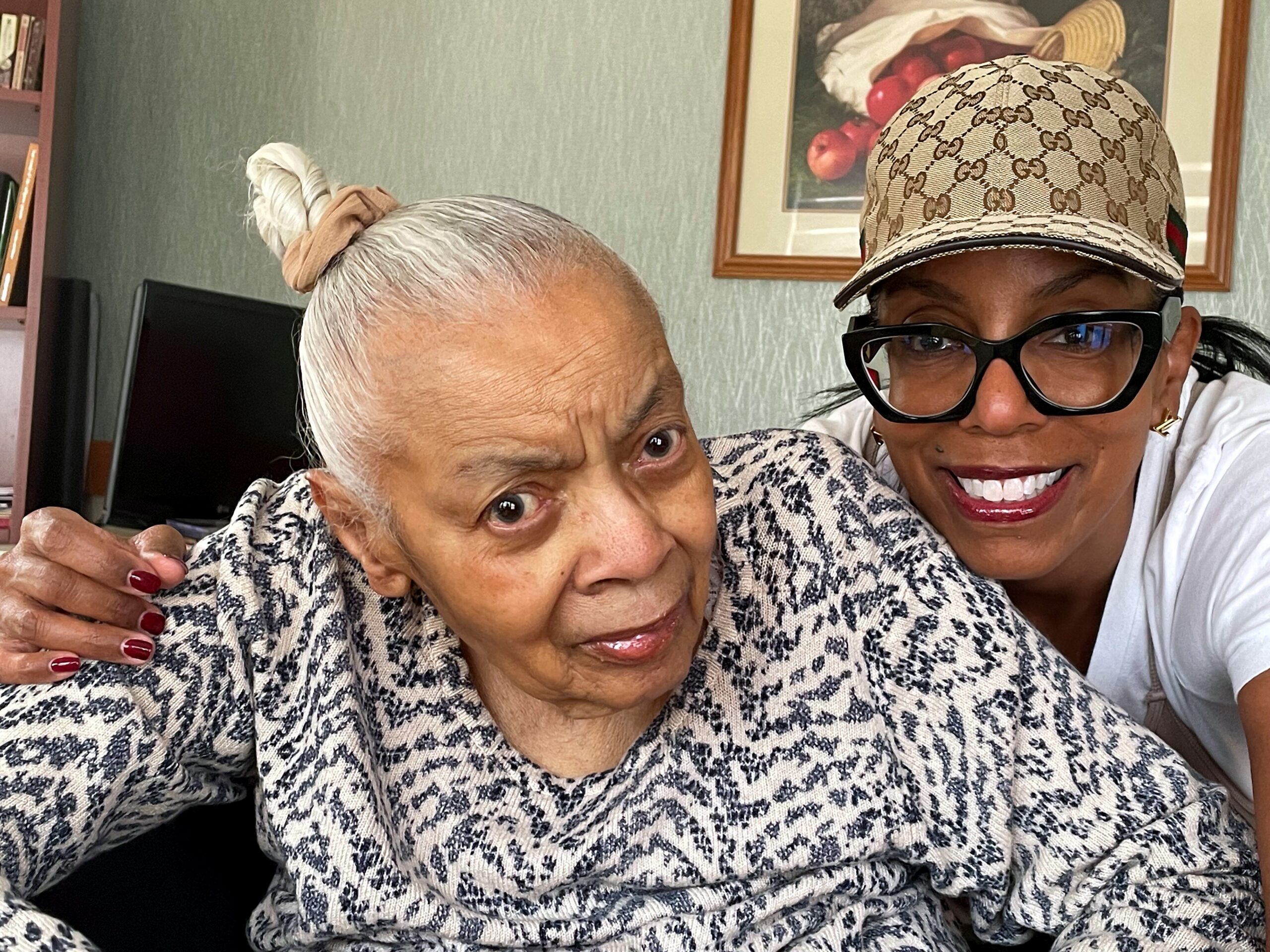 Two women taking a selfie indoors; one has a white hair bun and a patterned cardigan, the other wears a Gucci cap and black glasses, both smiling.