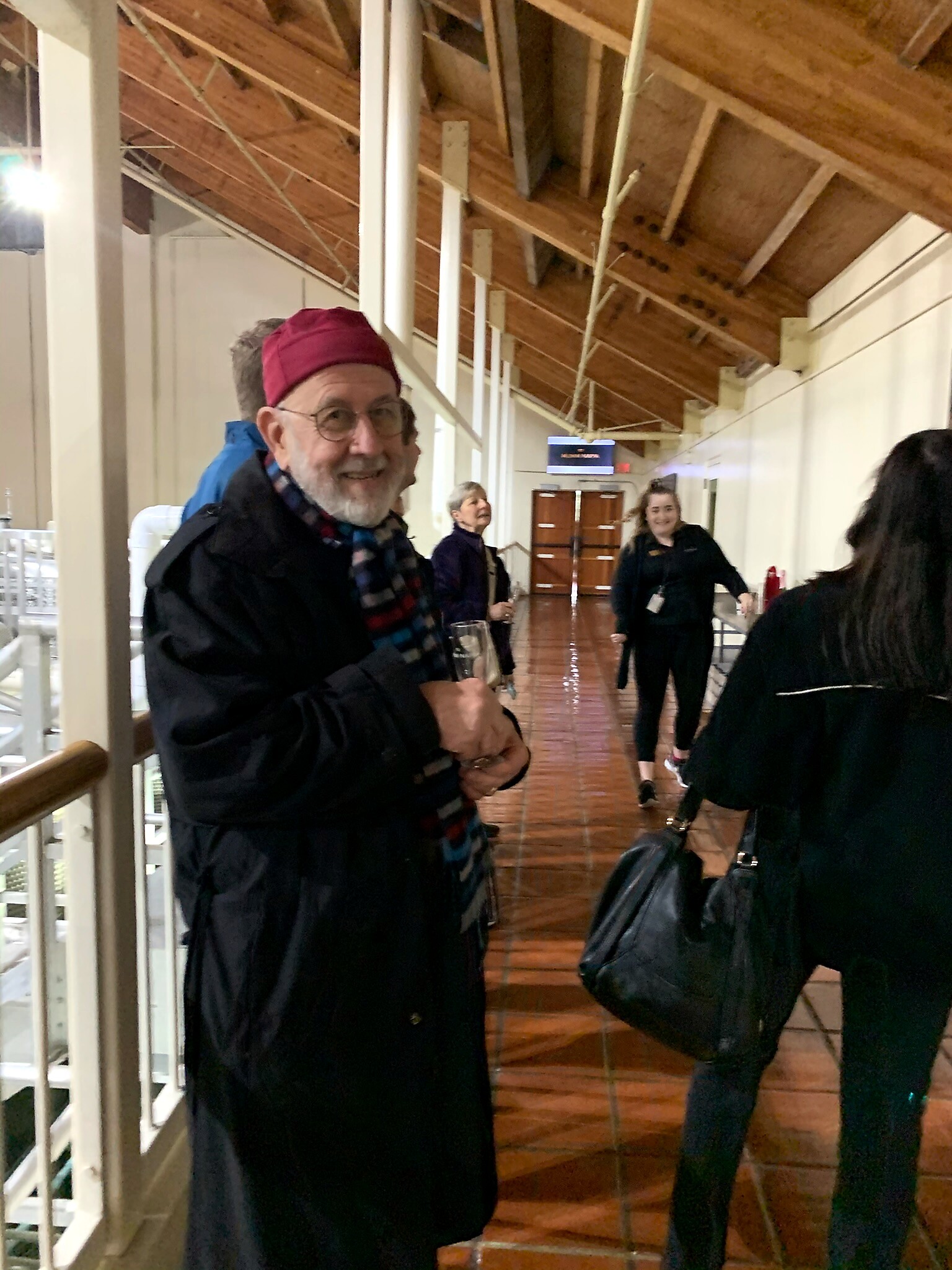 Smiling older man wearing a red beret and plaid scarf in a hallway with wooden beams, others in background.