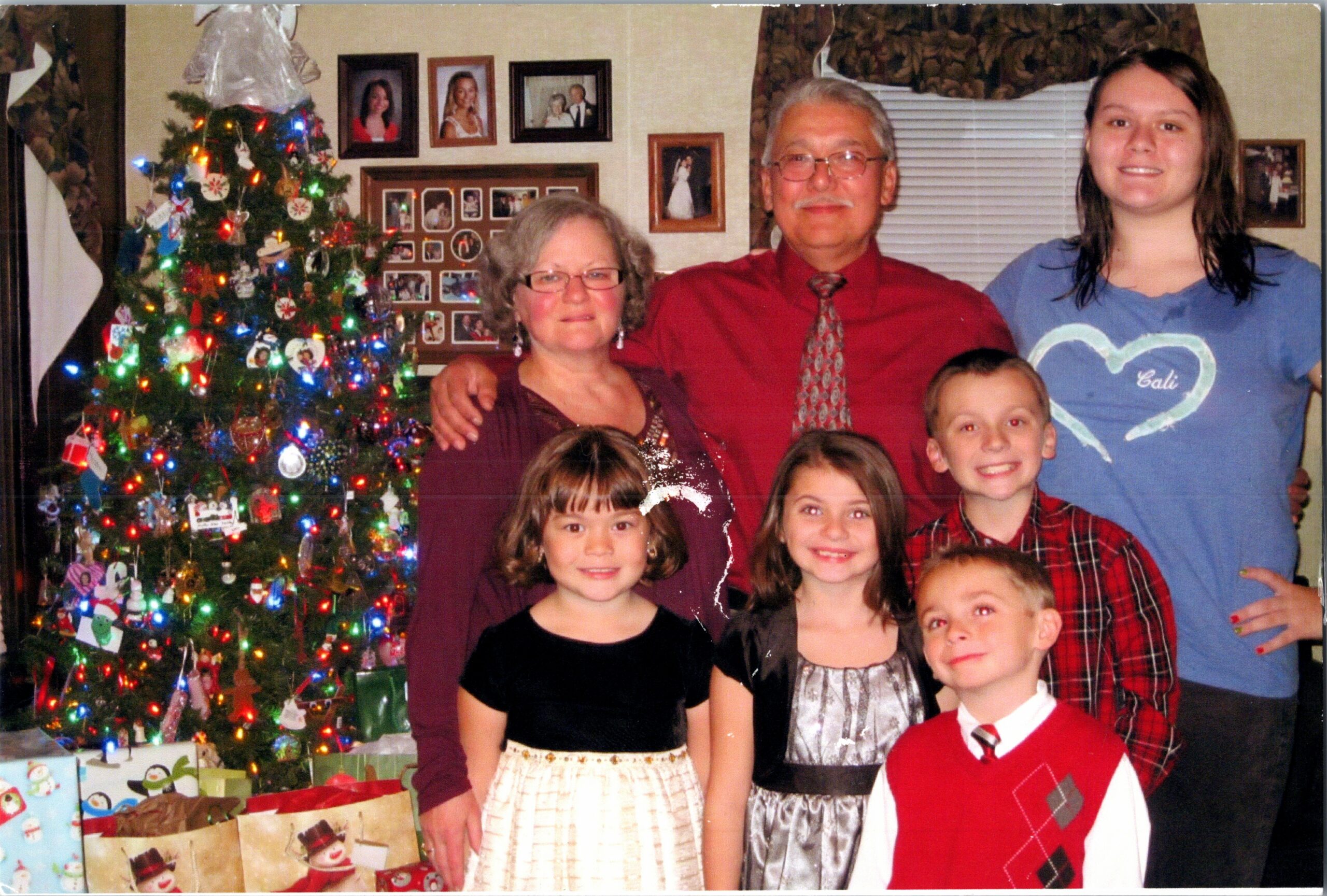 Family posing for a Christmas photo by a decorated tree with presents on the floor in a living room.