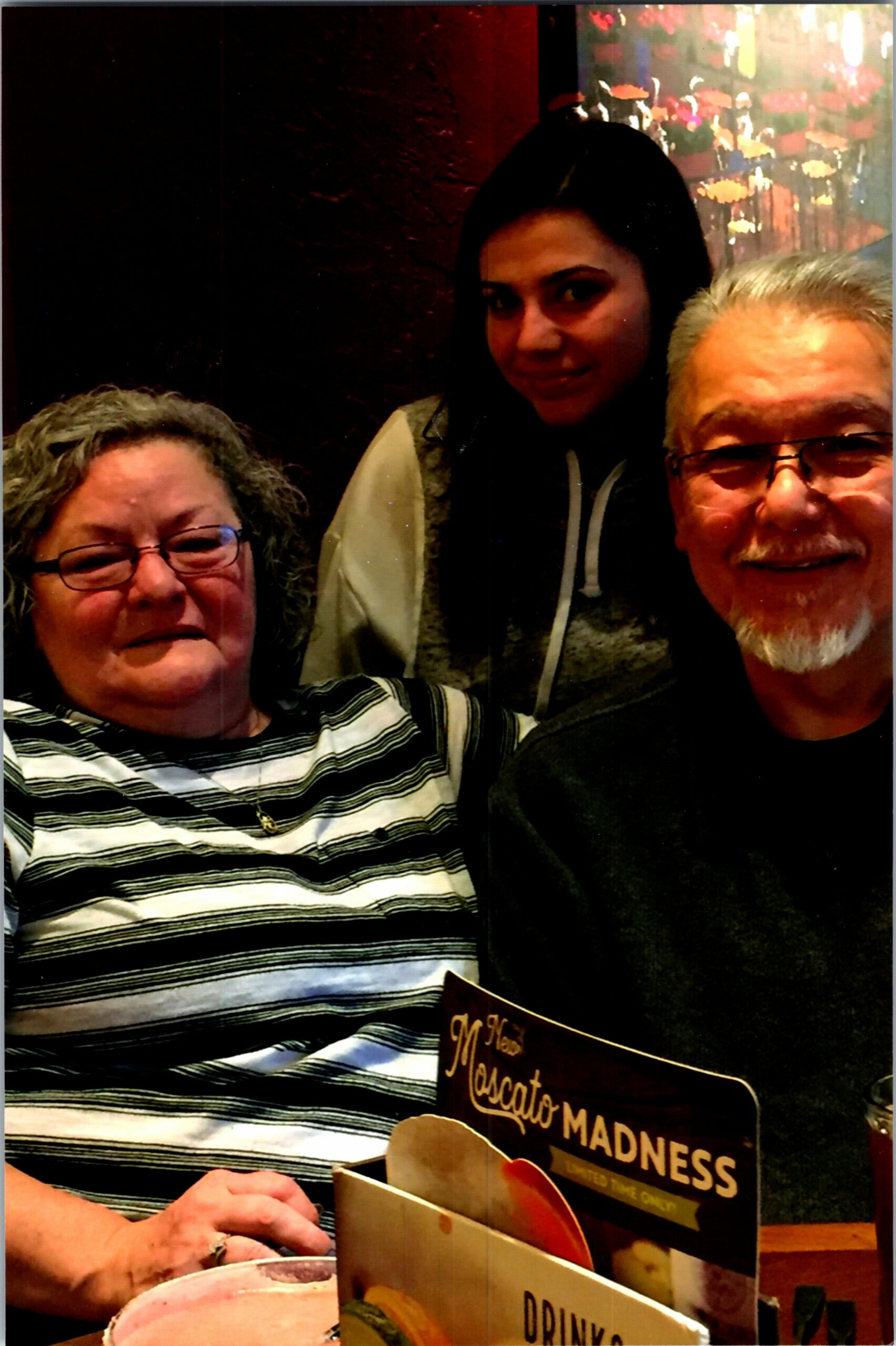 Three people pose for a photo in a restaurant; an older woman with glasses on the left, a younger woman in the middle, and a smiling man on the right, with menus visible in the foreground.