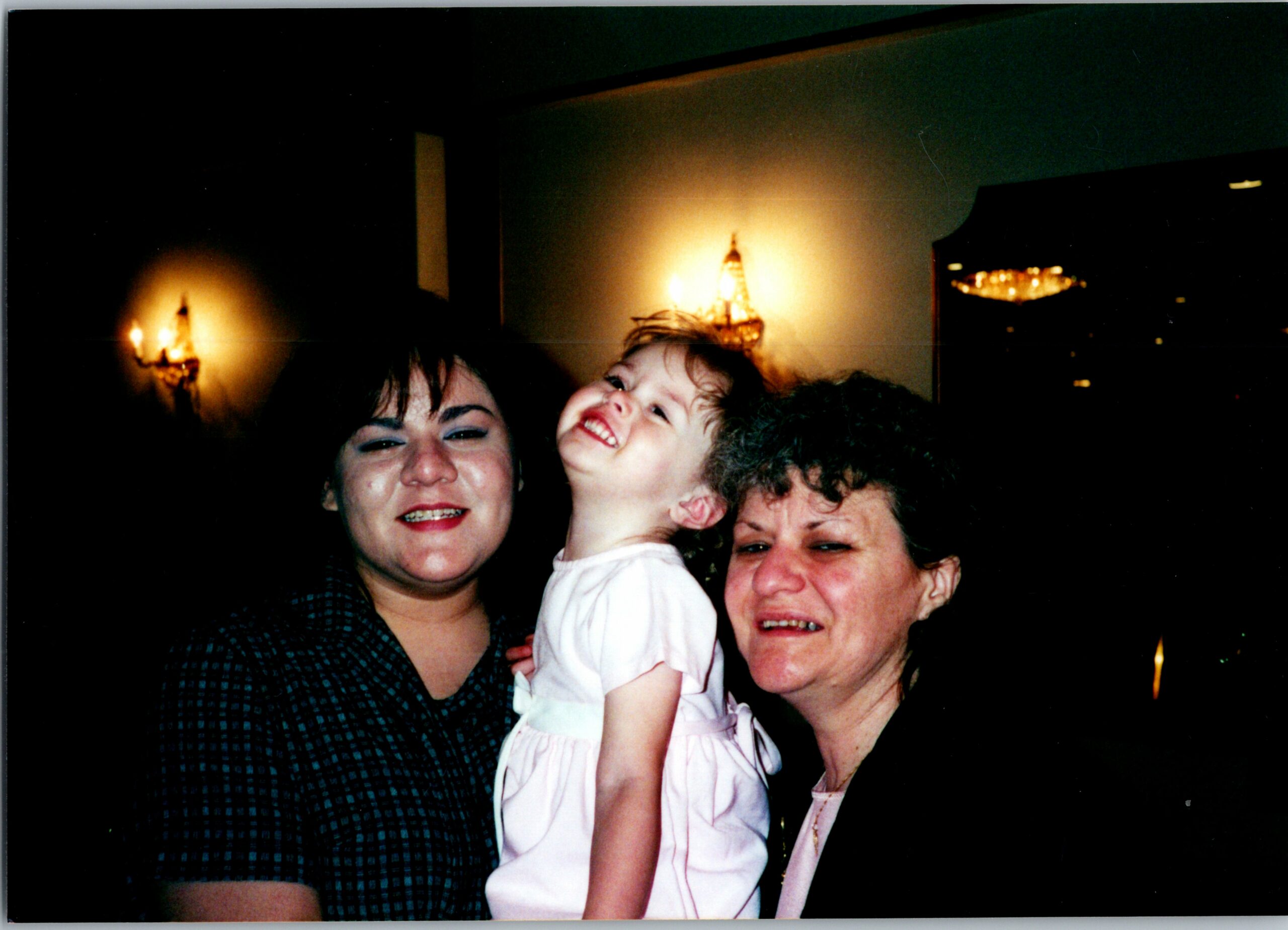 Three smiling people posing closely for a photo at a dimly lit indoor setting with a chandelier in the background.