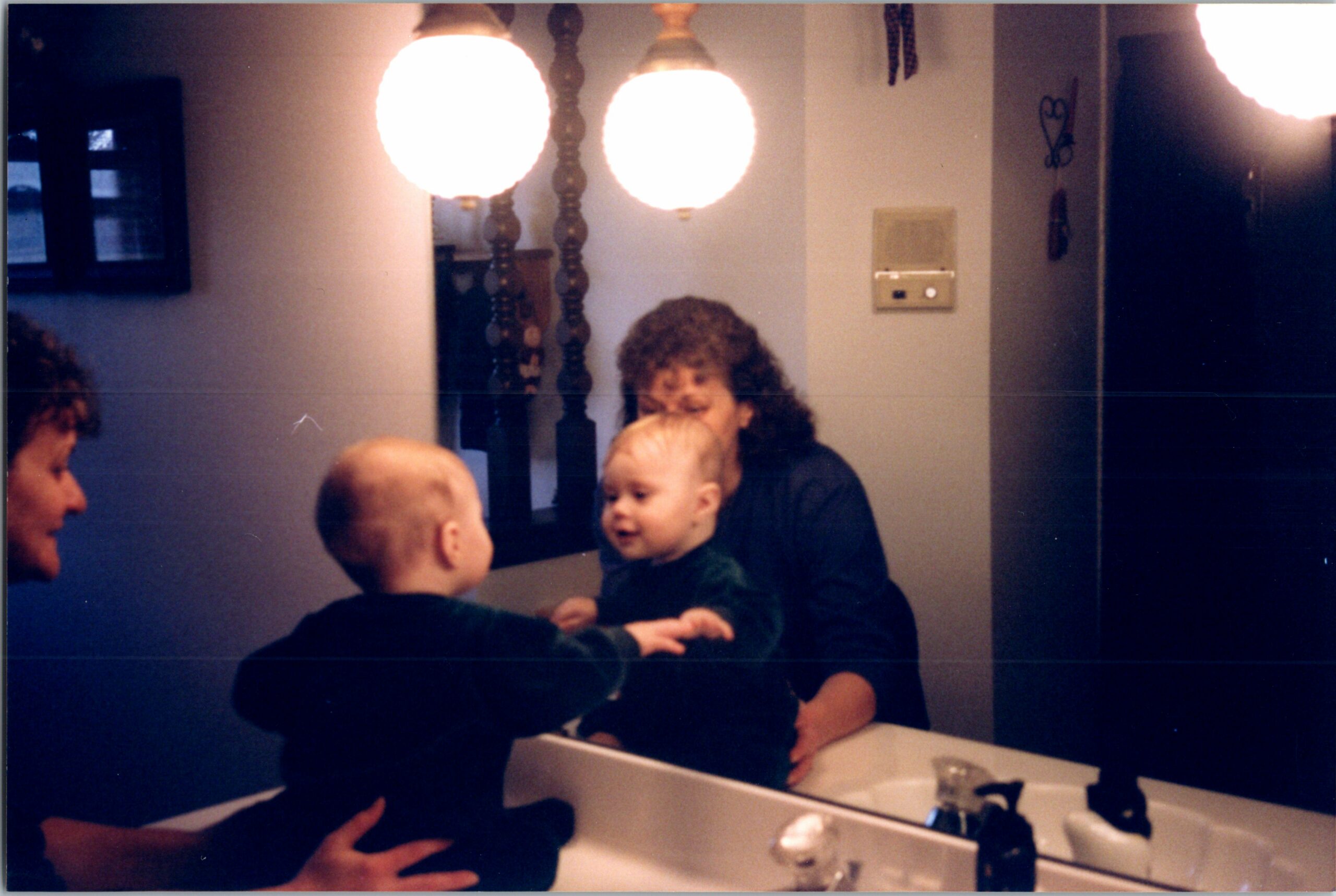 Family in a dim bathroom around the sink, with a baby facing forward and adults nearby seen in the mirror reflection.