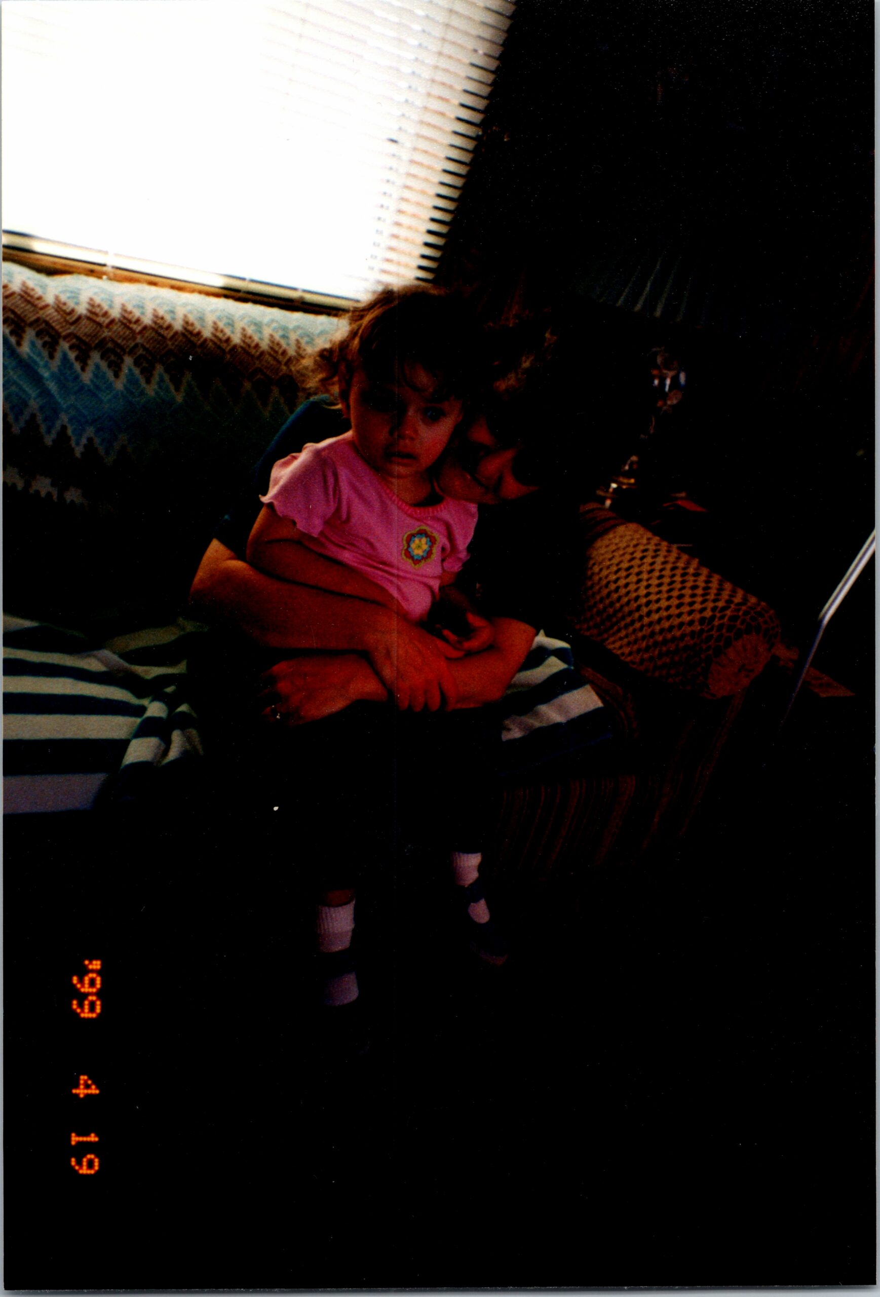 A young girl in a pink shirt sits on an adult's lap on a patterned sofa by a window with blinds.