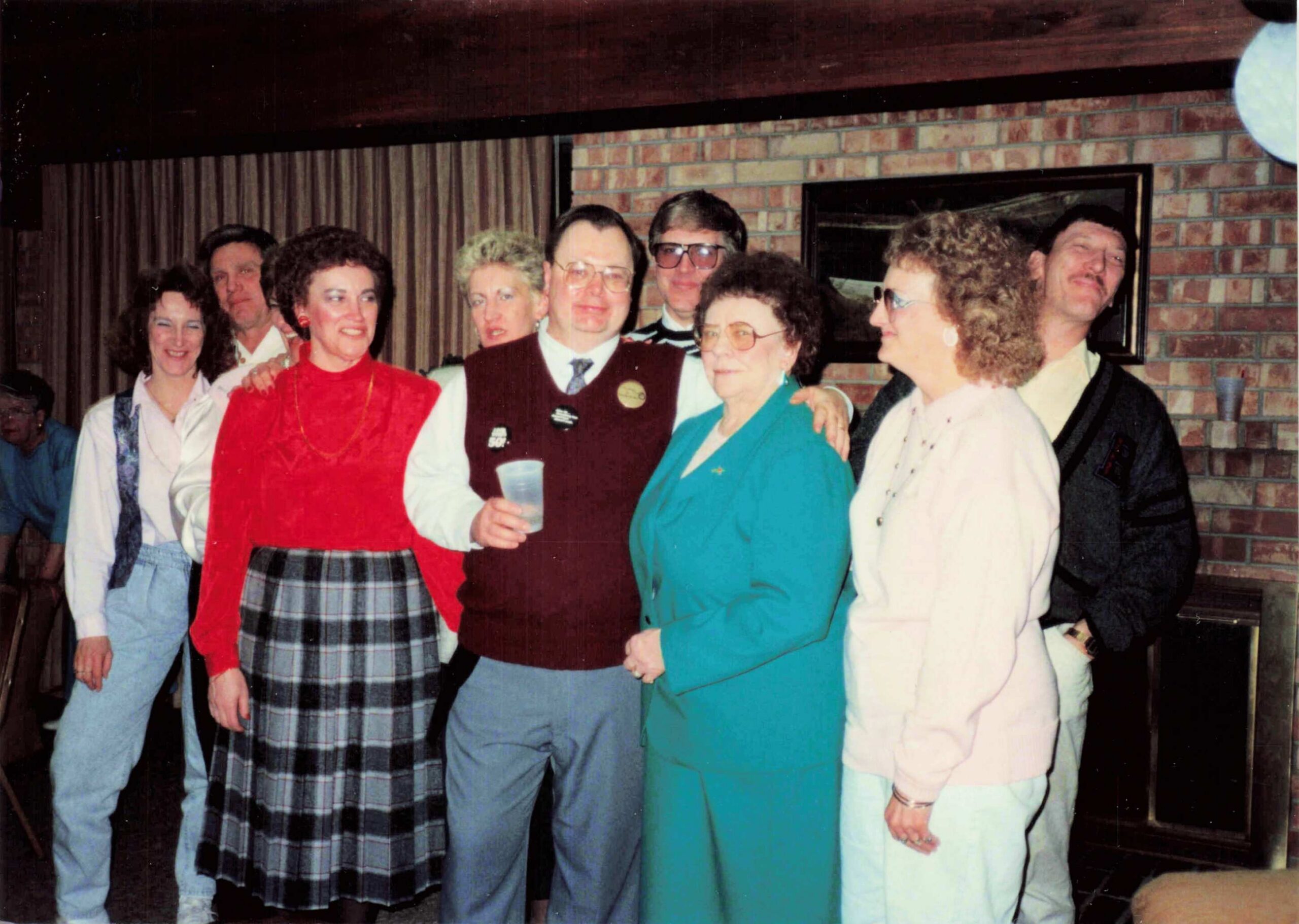 Group of smiling adults standing close together at a casual indoor gathering, wearing colorful 1980s-style clothing in front of a brick wall and curtains.