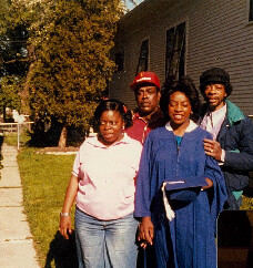 Group of four African American people posing outside a house; a graduate in a blue gown and cap holds a diploma, flanked by two women and a man smiling.