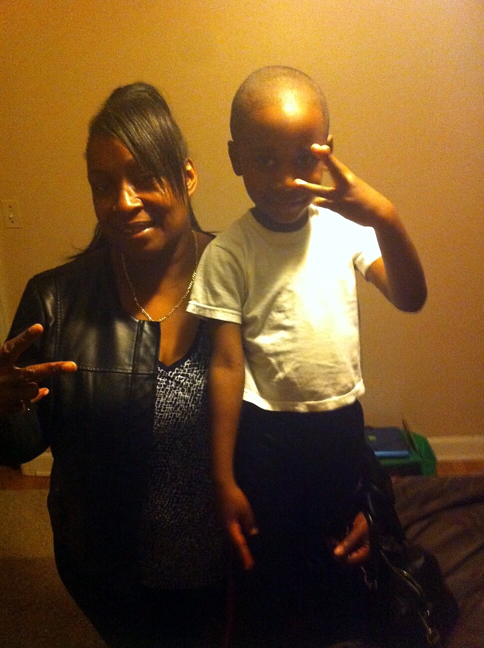 Woman and child posing together indoors, both making peace signs toward the camera on a beige wall backdrop.