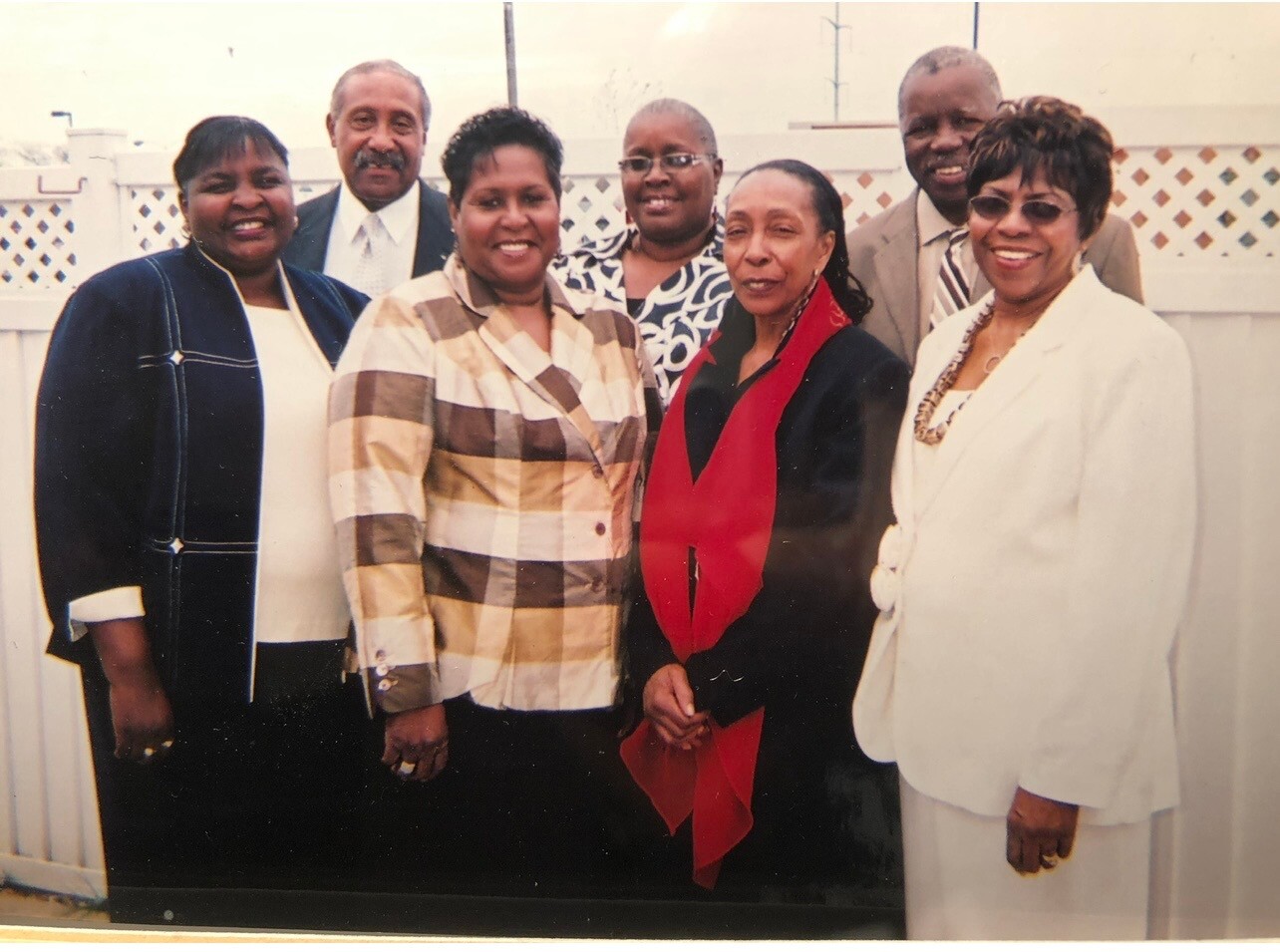 Seven adults pose outdoors in front of a white lattice fence, dressed in business and smart-casual attire, smiling at the camera.