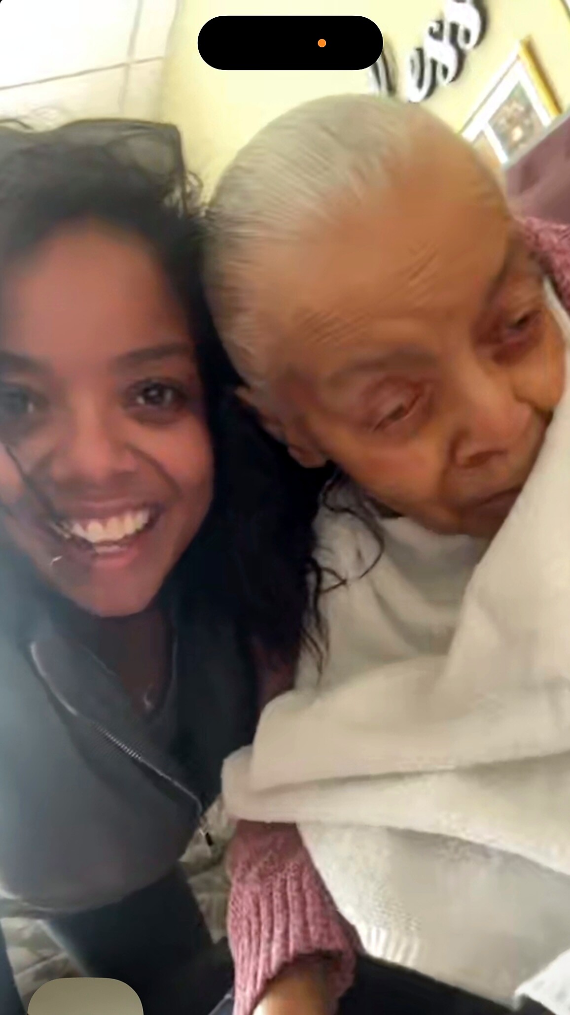 Two people pose for a close selfie: a smiling woman on the left and an elderly person on the right indoors, cuddled together.