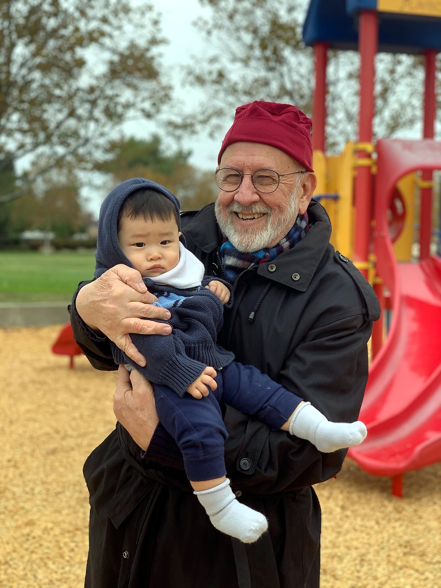 Grandfather in a red hat tenderly holding a baby in a navy hoodie at a colorful playground.