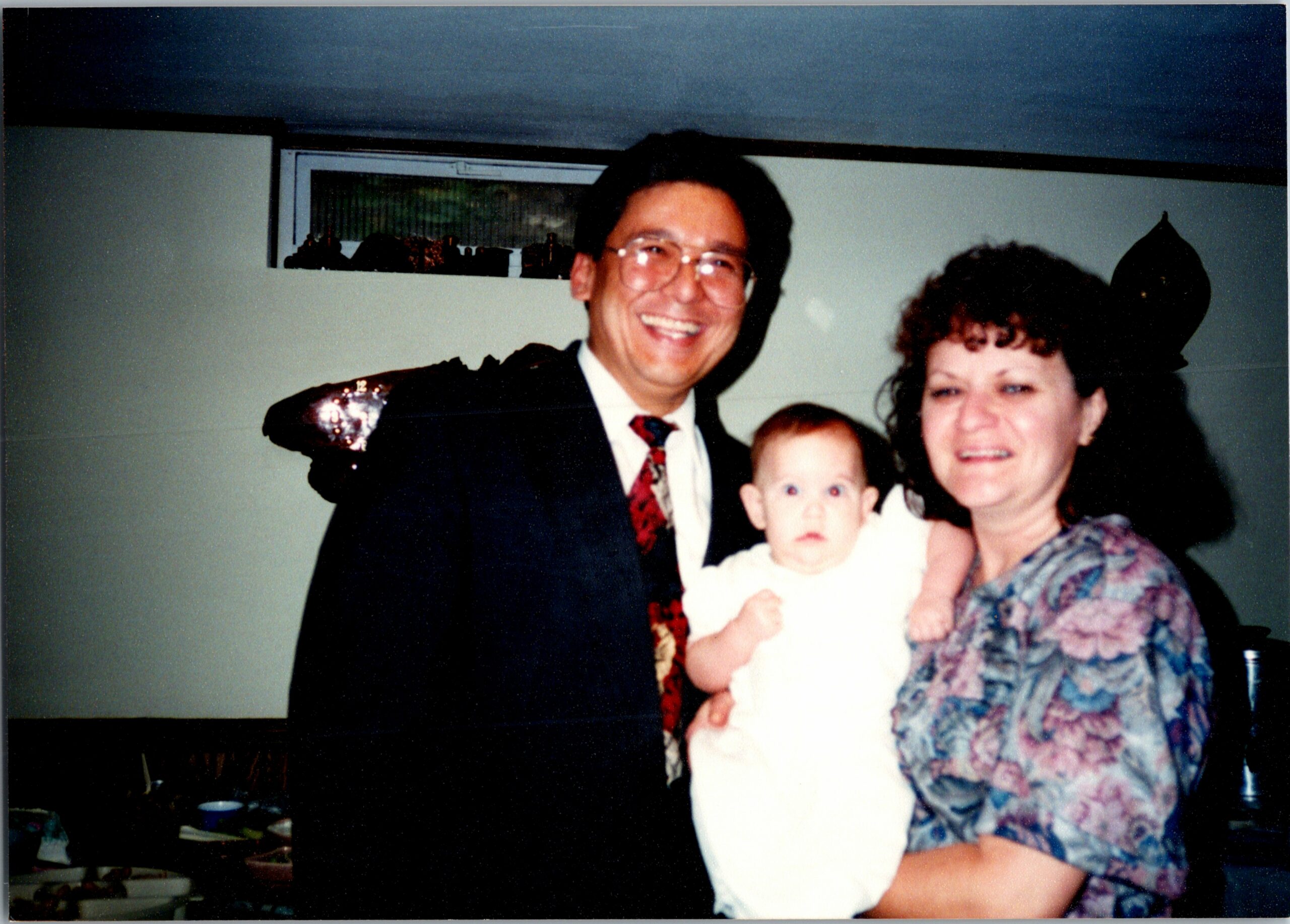 Smiling man in a suit holding a baby with a woman, indoors at a family gathering.