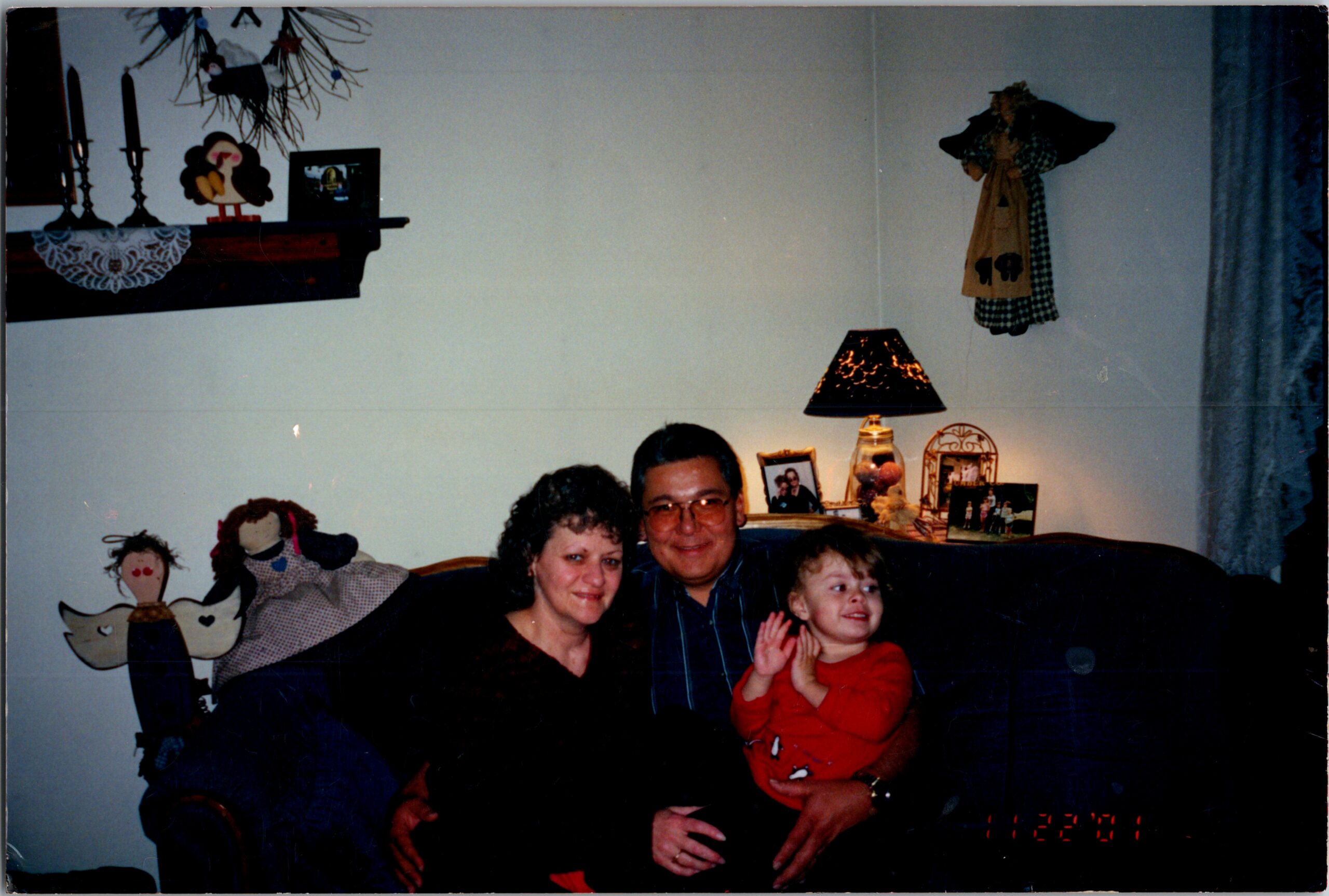 Family sits on a sofa in a living room; a child in red sits with adults, with photos and decorations on a shelf behind them.