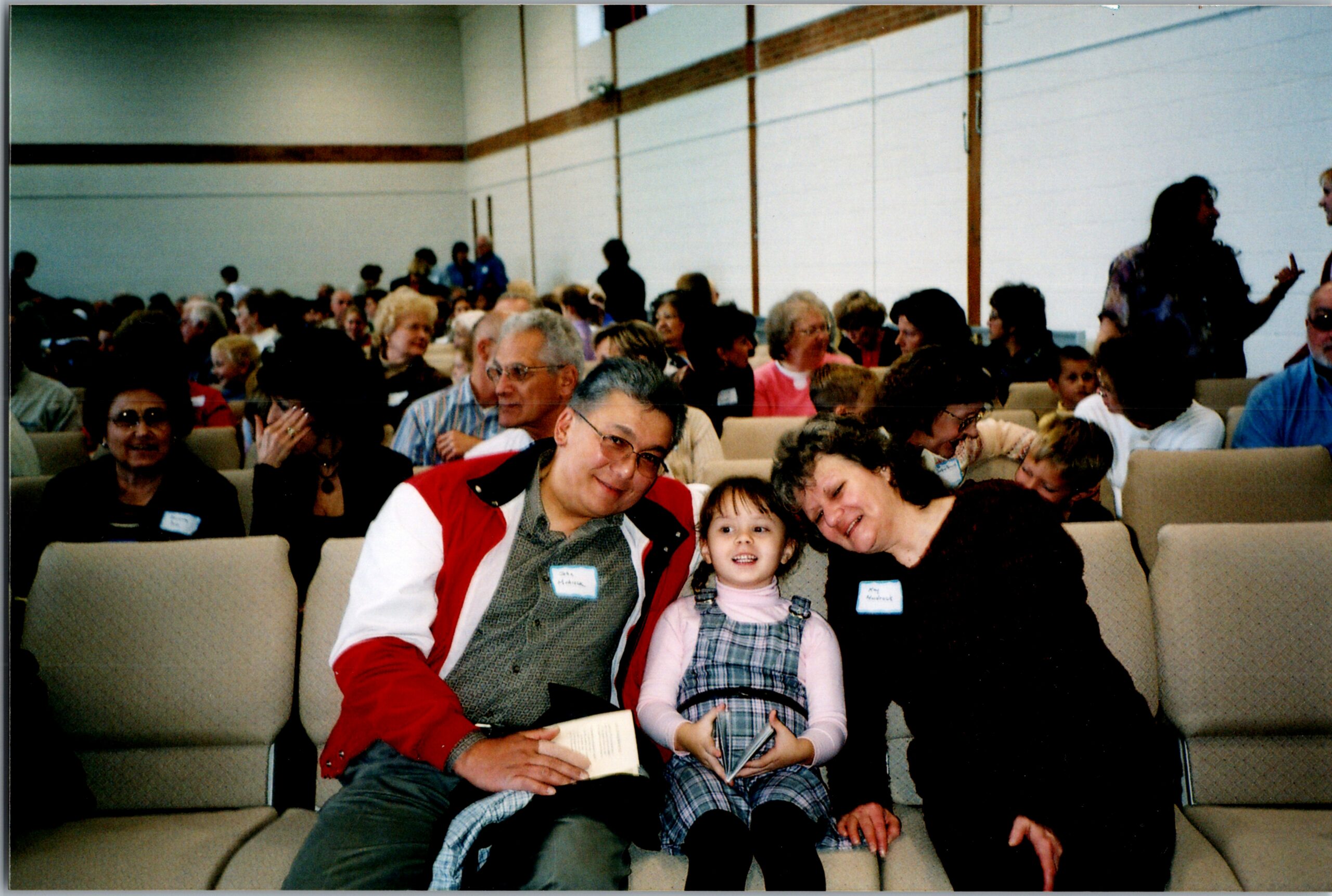 Family and friends sit in rows of chairs at a community event; foreground shows a man in a red jacket with a young girl in a plaid dress smiling at the camera.