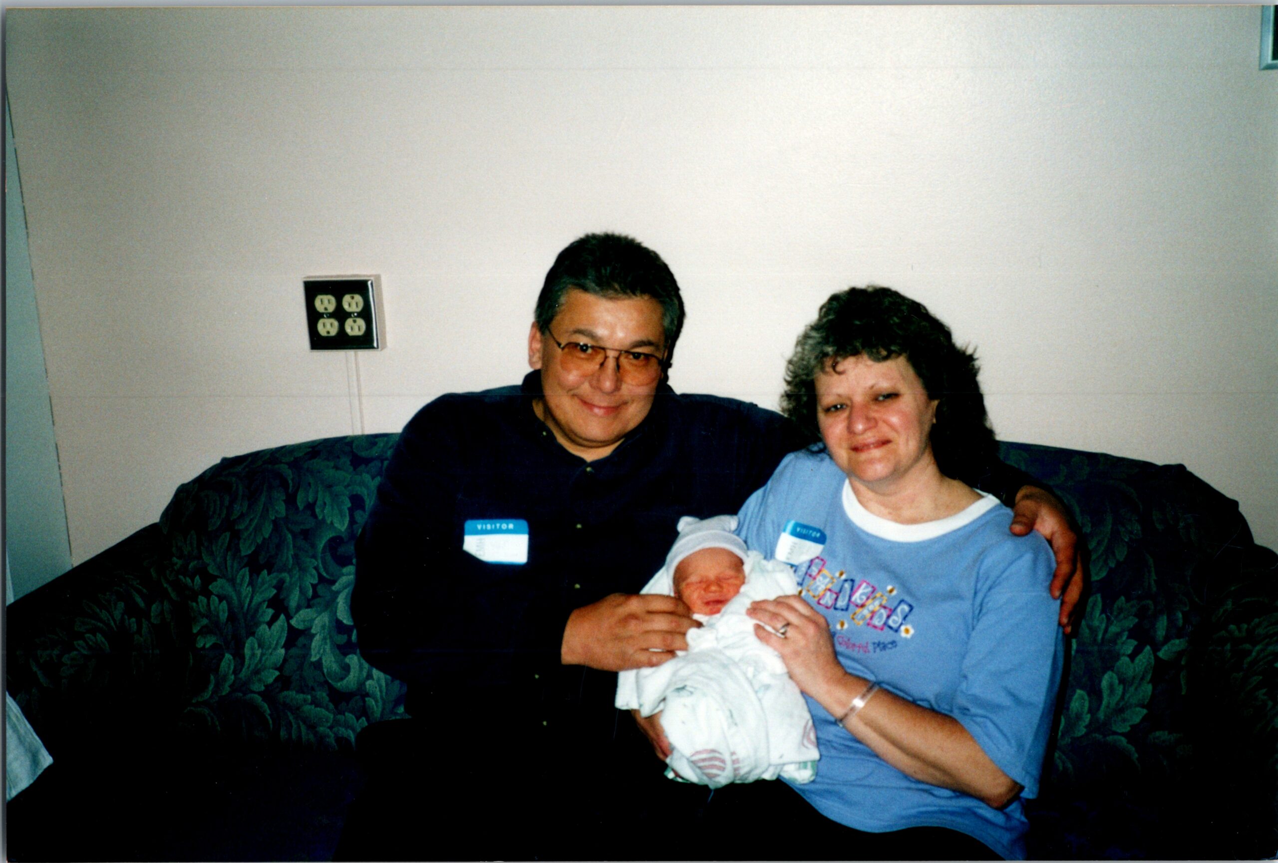 Smiling couple on a patterned sofa cradle a newborn baby wrapped in a white blanket.