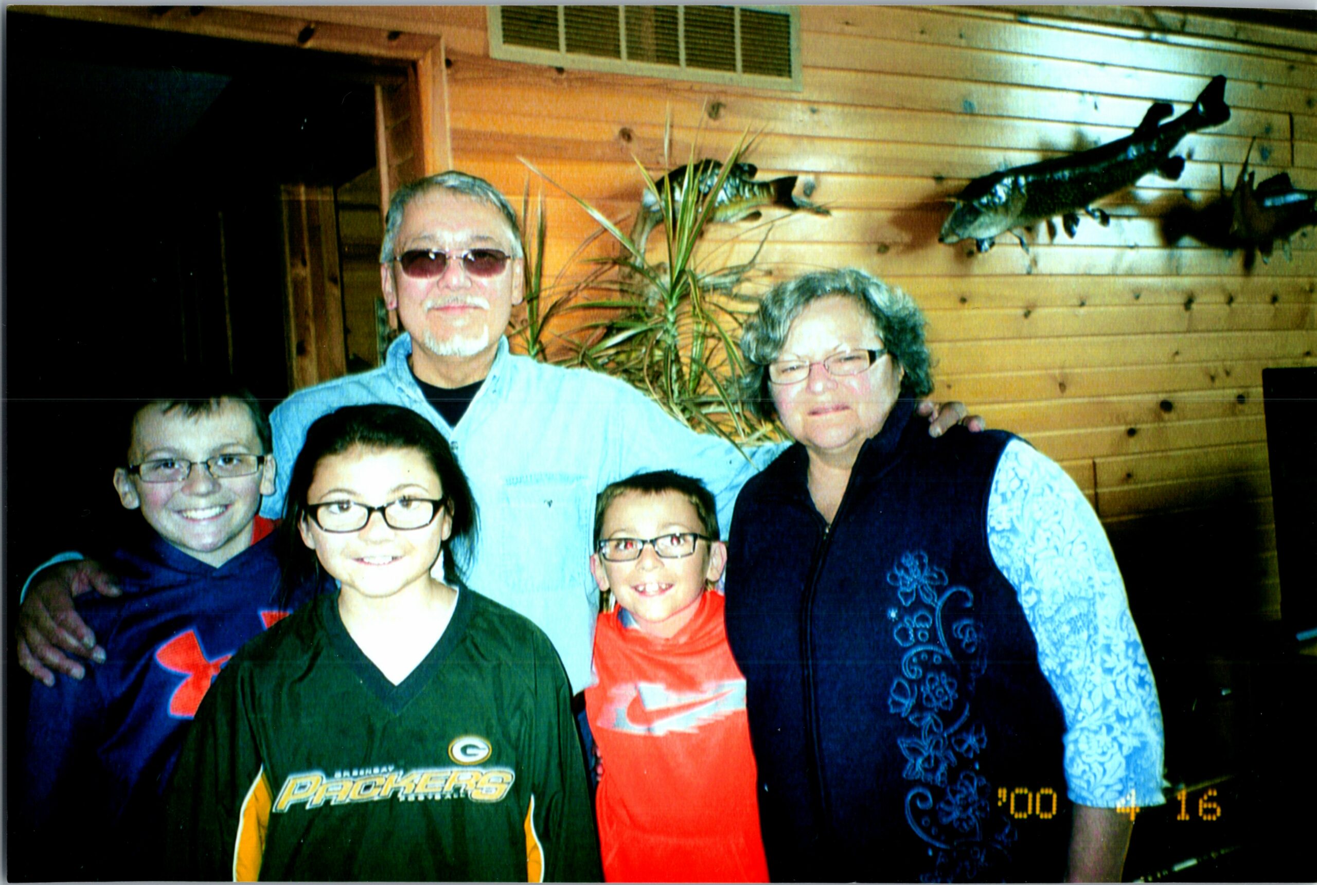 Group portrait of six family members posing indoors against a wood-paneled wall, with three children in front and two adults behind, all smiling at the camera.