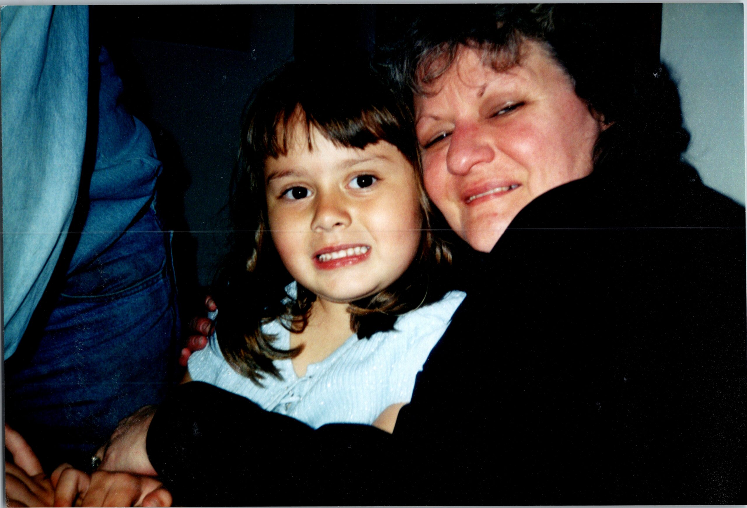 A young girl with light brown hair smiling beside an adult woman who is also smiling, close together in a dark setting.