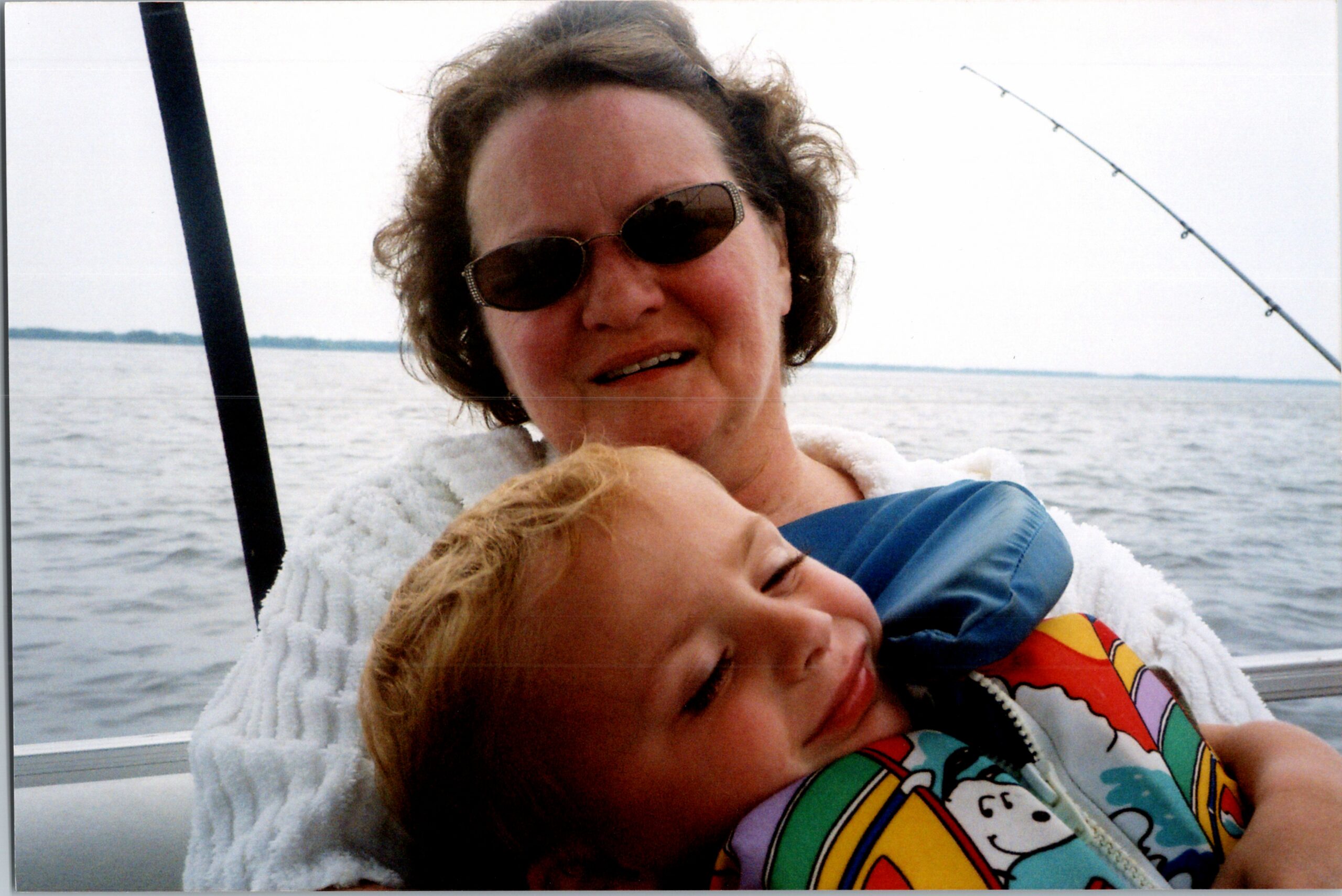 Two people hugging on a boat with water in the background; woman wearing sunglasses and a white fleece, child in a colorful life jacket smiles into the embrace.