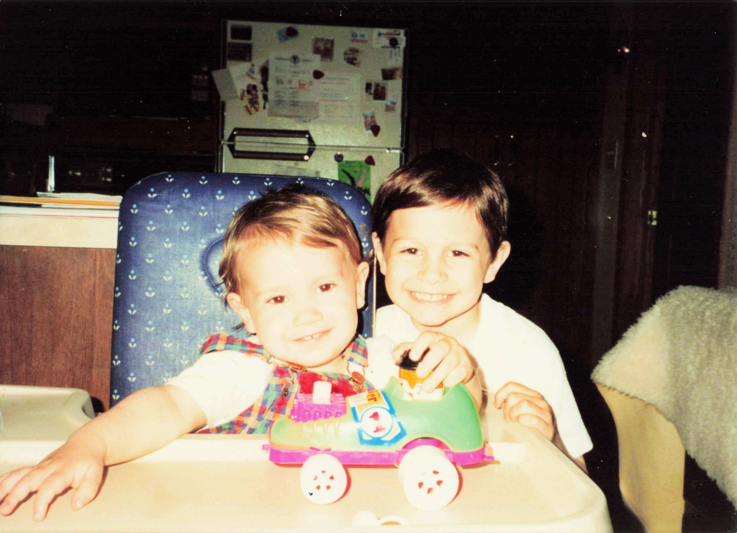 Two young children smile at the camera while a bright toy car sits on a table in front of them, fridge with magnets in the background.