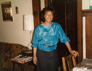 Woman in a blue satin blouse stands in a living room, resting hand on a wooden chair and smiling at the camera.