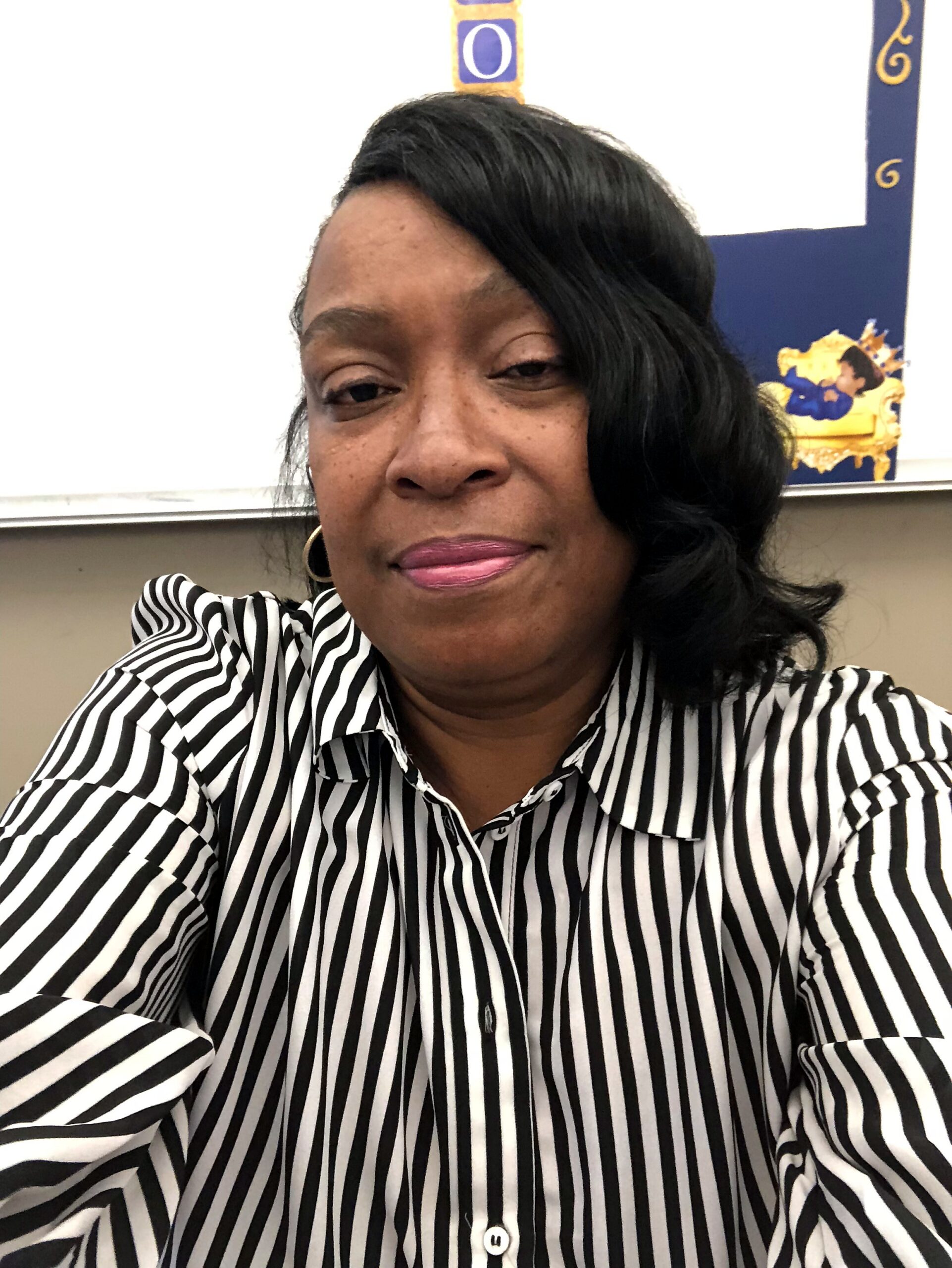 Close-up selfie of a woman wearing a black-and-white striped shirt and gold hoop earrings in a classroom setting, smiling slightly.