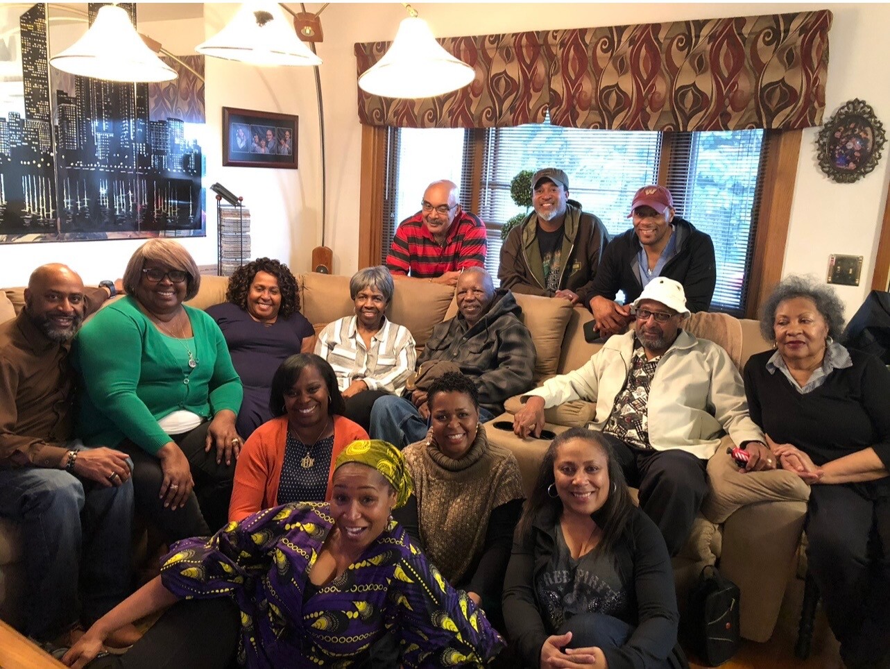 Group of 14 adults posing for a photo on a beige couch and floor in a living room, smiling at the camera.