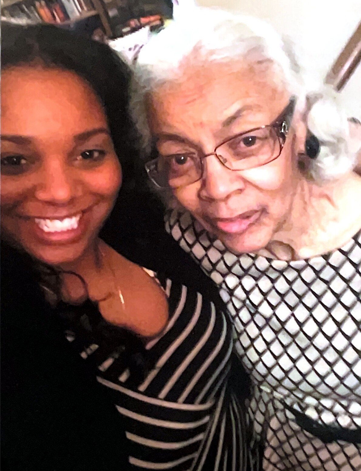Two women smile for a selfie: a young Black woman in a striped top beside an older white-haired woman with glasses indoors.