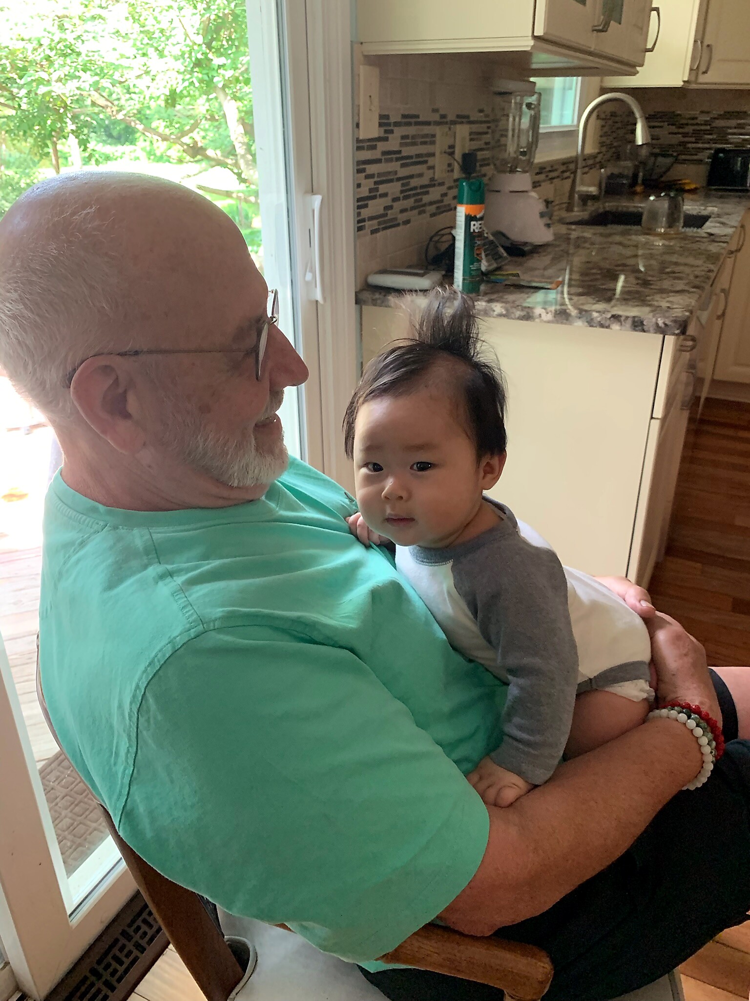Elderly man with glasses and a white beard holding a young child in a kitchen by a window and blender on the counter.