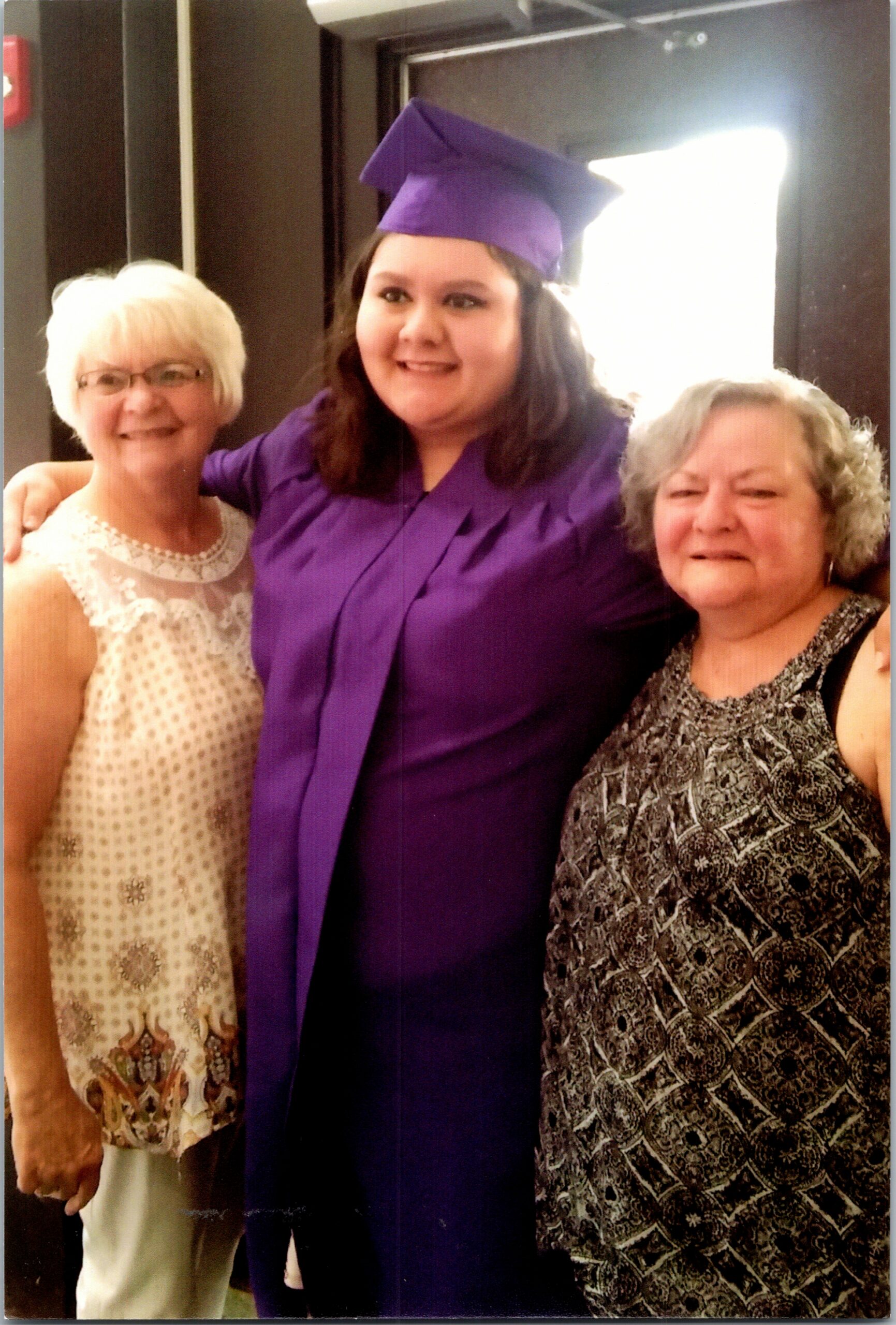 Three generations celebrate: a young graduate in a purple cap and gown stands with two smiling older women indoors, one in a light patterned sleeveless top and the other in a black-and-white patterned dress.