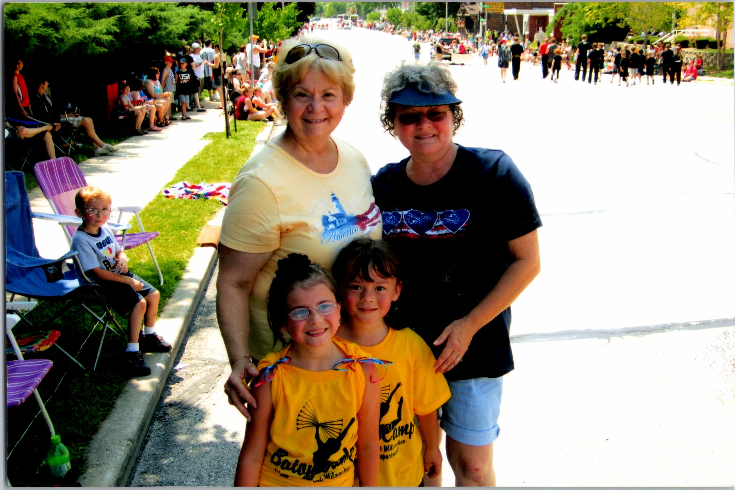 Two women and three children smile at the camera in the foreground as a sunny parade route fills with spectators along the curb.