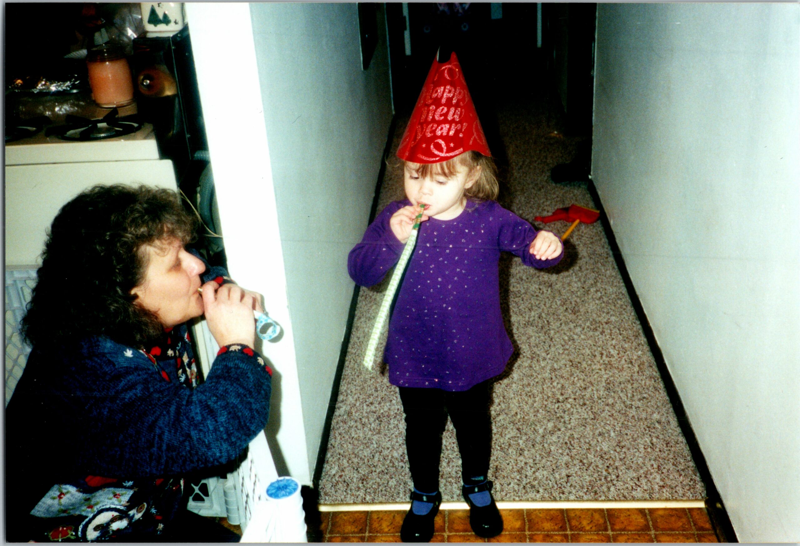 Young child in a red 'Happy New Year' party hat blowing a party horn in a home hallway, with an adult nearby.